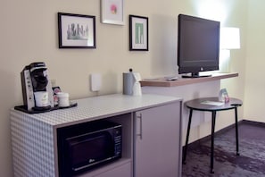 A hotel room with a kitchenette, a flat-screen TV, framed artwork on the wall, and a small table with a lamp.
