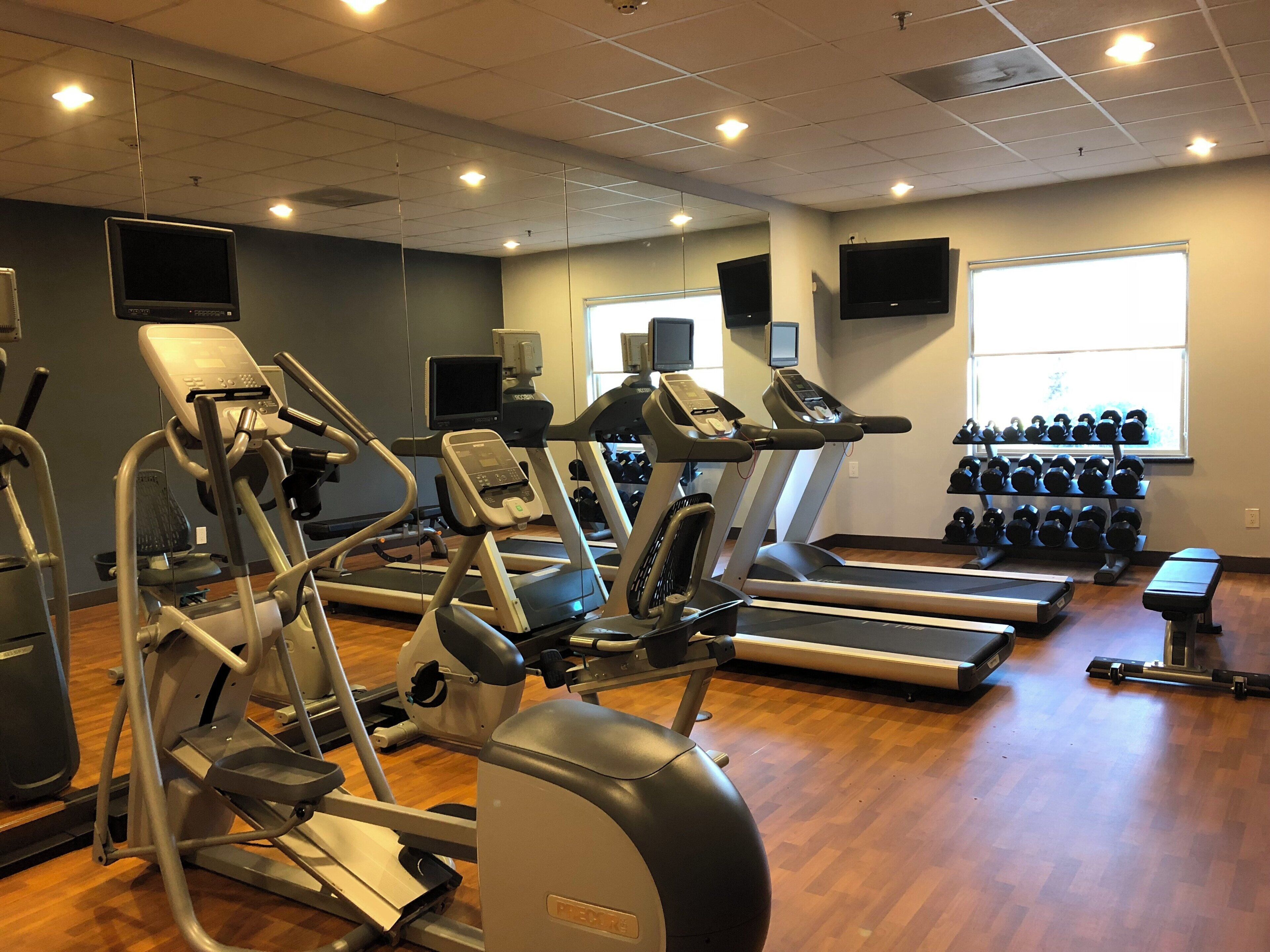 Ein Fitnessstudio mit verschiedenen Trainingsgeräten, einem an der Wand montierten Fernseher und einem Fenster mit Tageslicht.