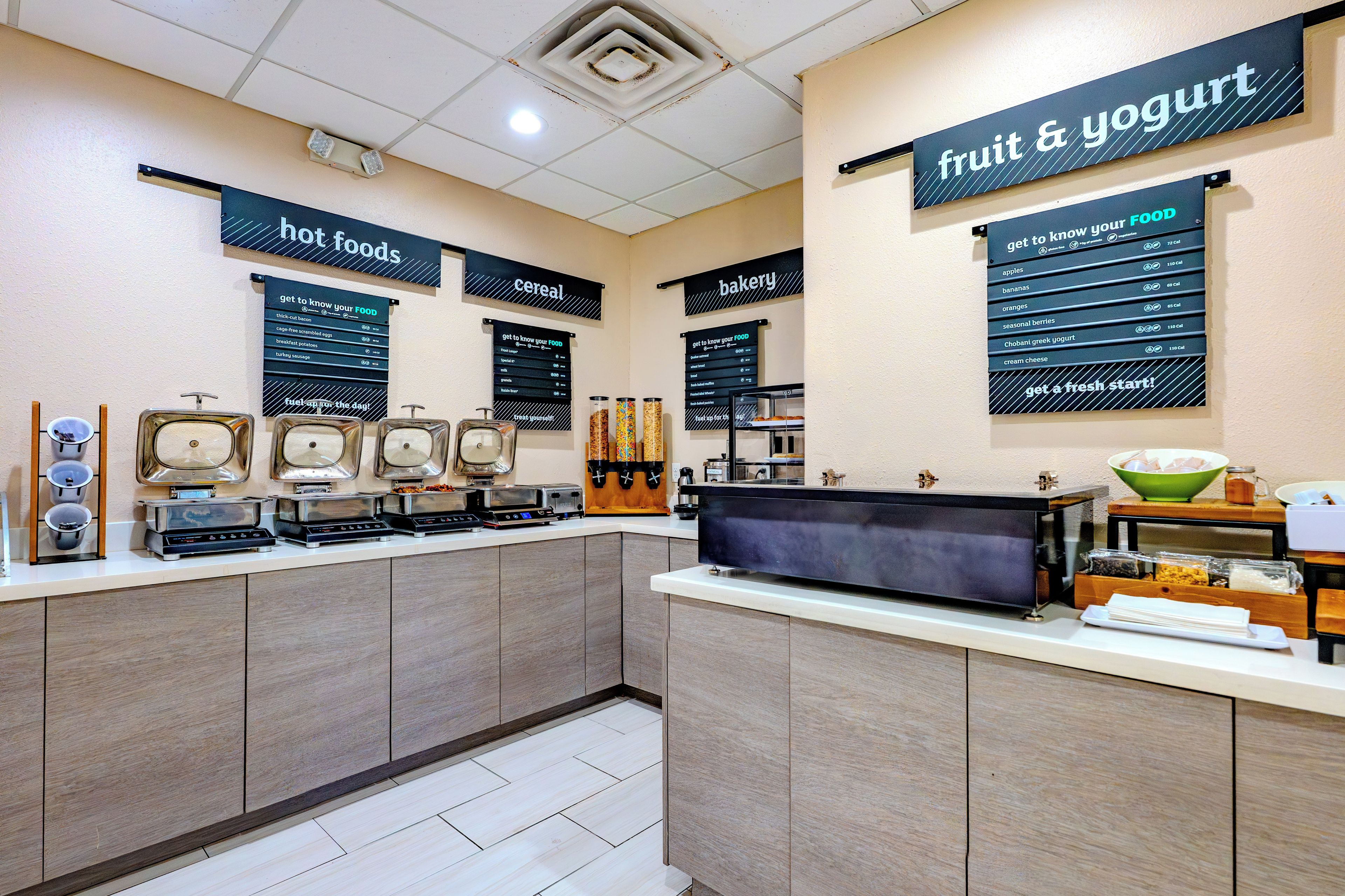 A self-service food station with labeled dispensers for hot foods, cereal, bakery items, and fruit & yogurt.