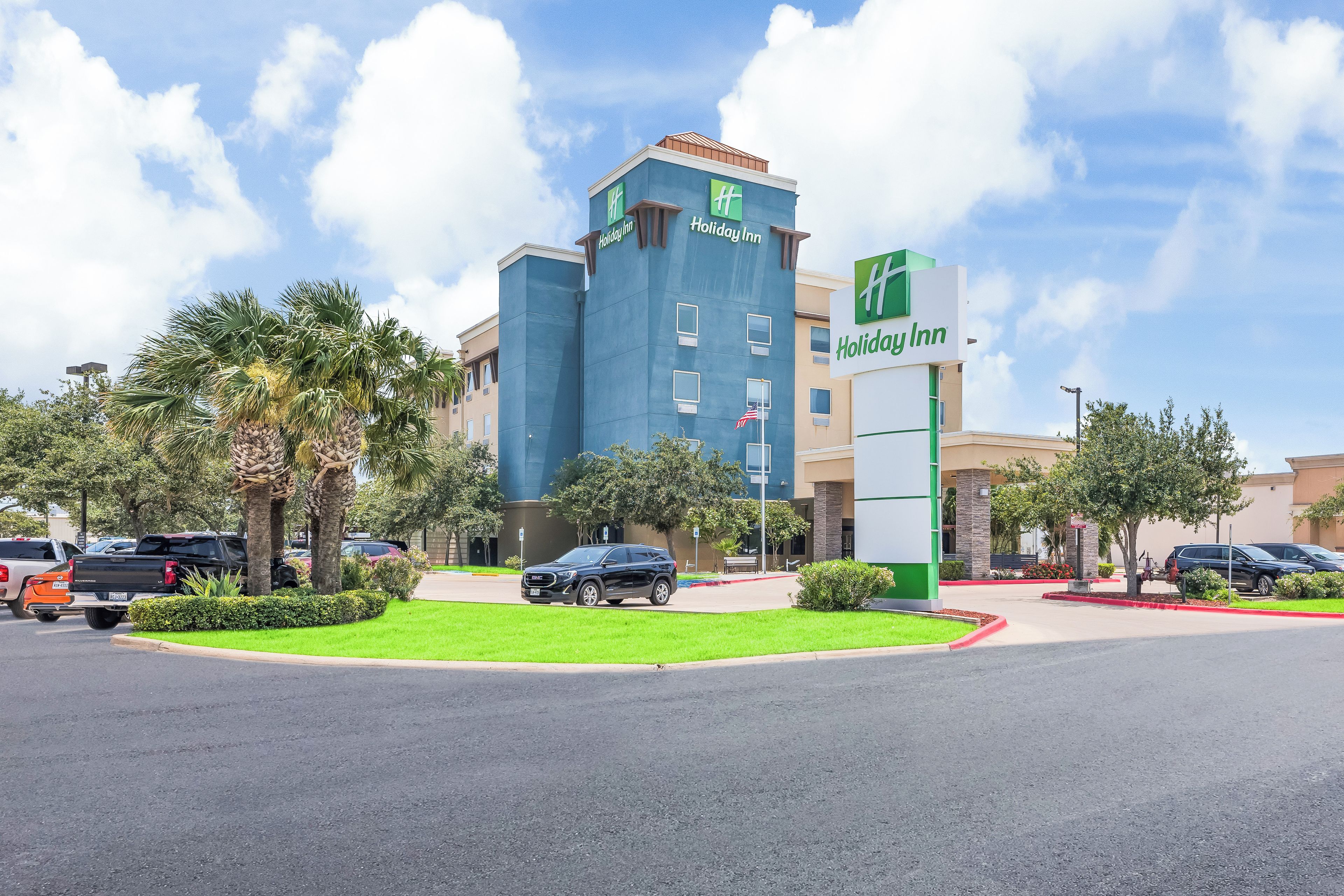 Un hotel Holiday Inn con fachada azul, palmeras y un estacionamiento con coches.