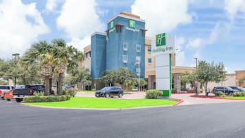 Un hotel Holiday Inn con fachada azul, palmeras y un estacionamiento con coches.