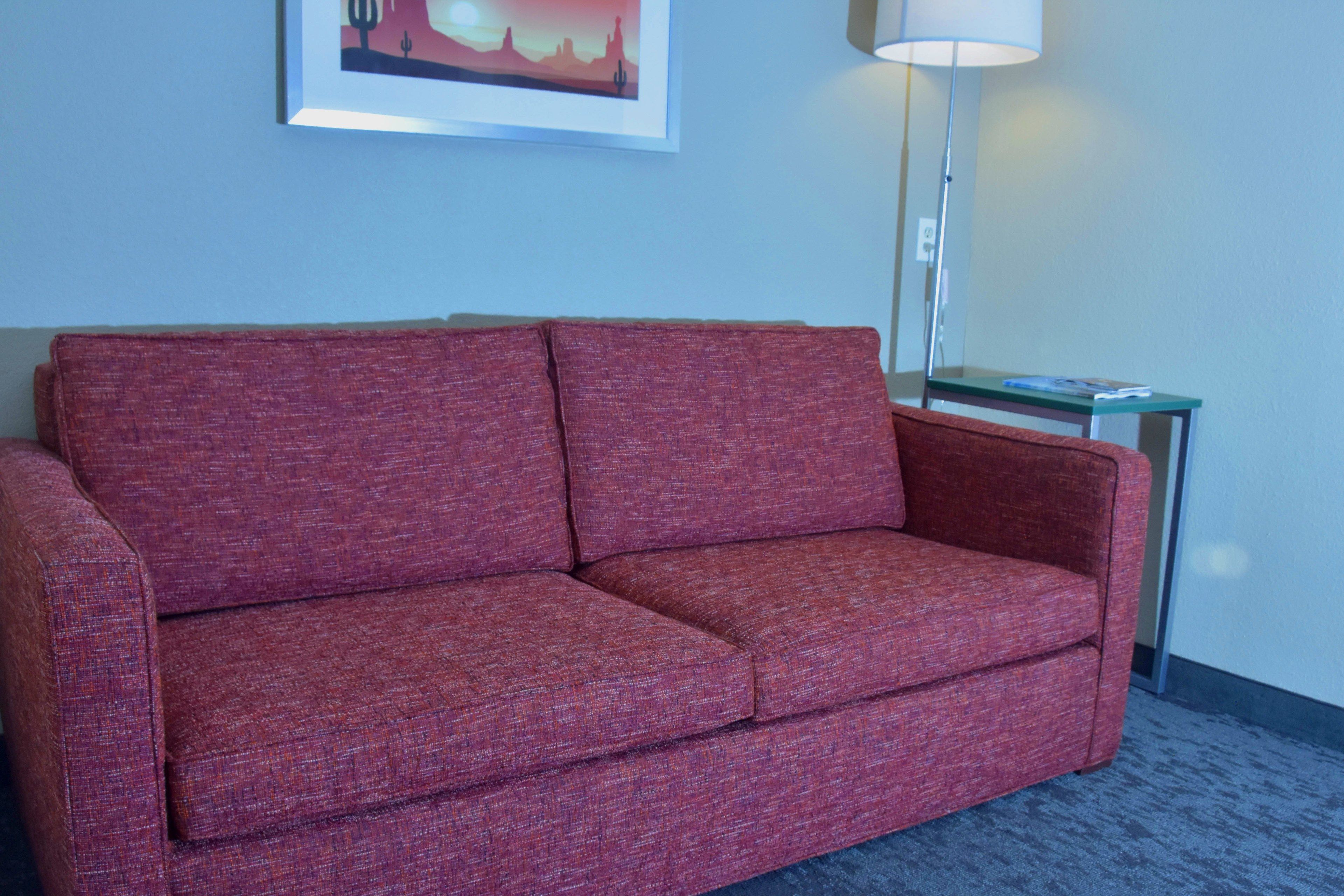 A red sofa in a room with a framed picture on the wall and a lamp on a side table.