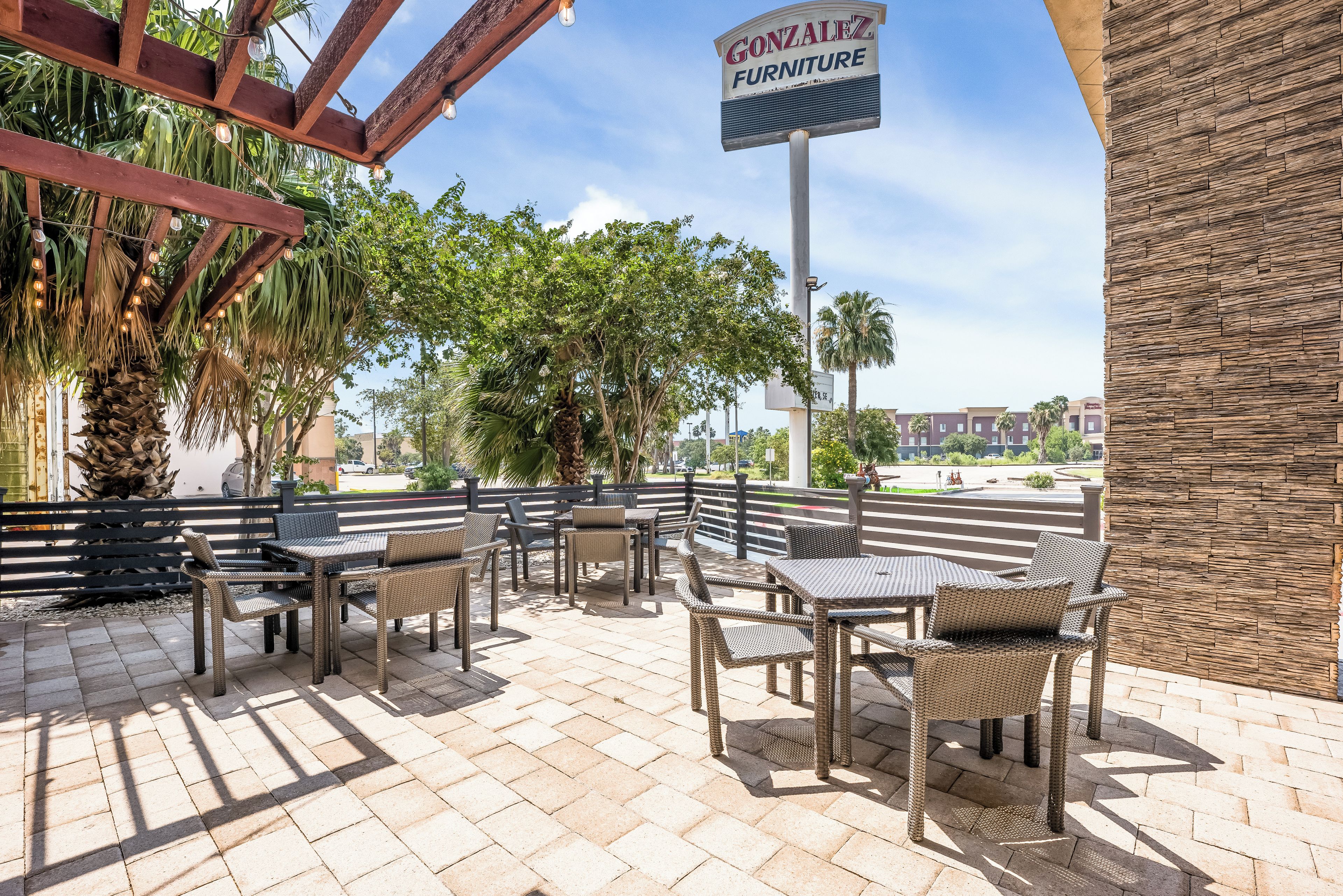 An outdoor seating area with wicker furniture, a sign for Gonzalez Furniture, and a view of a parking lot and buildings in the background.