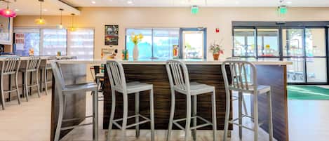 Un interior de restaurante con una zona de bar, diferentes distribuciones de mesas y vistas al exterior a través de las ventanas.