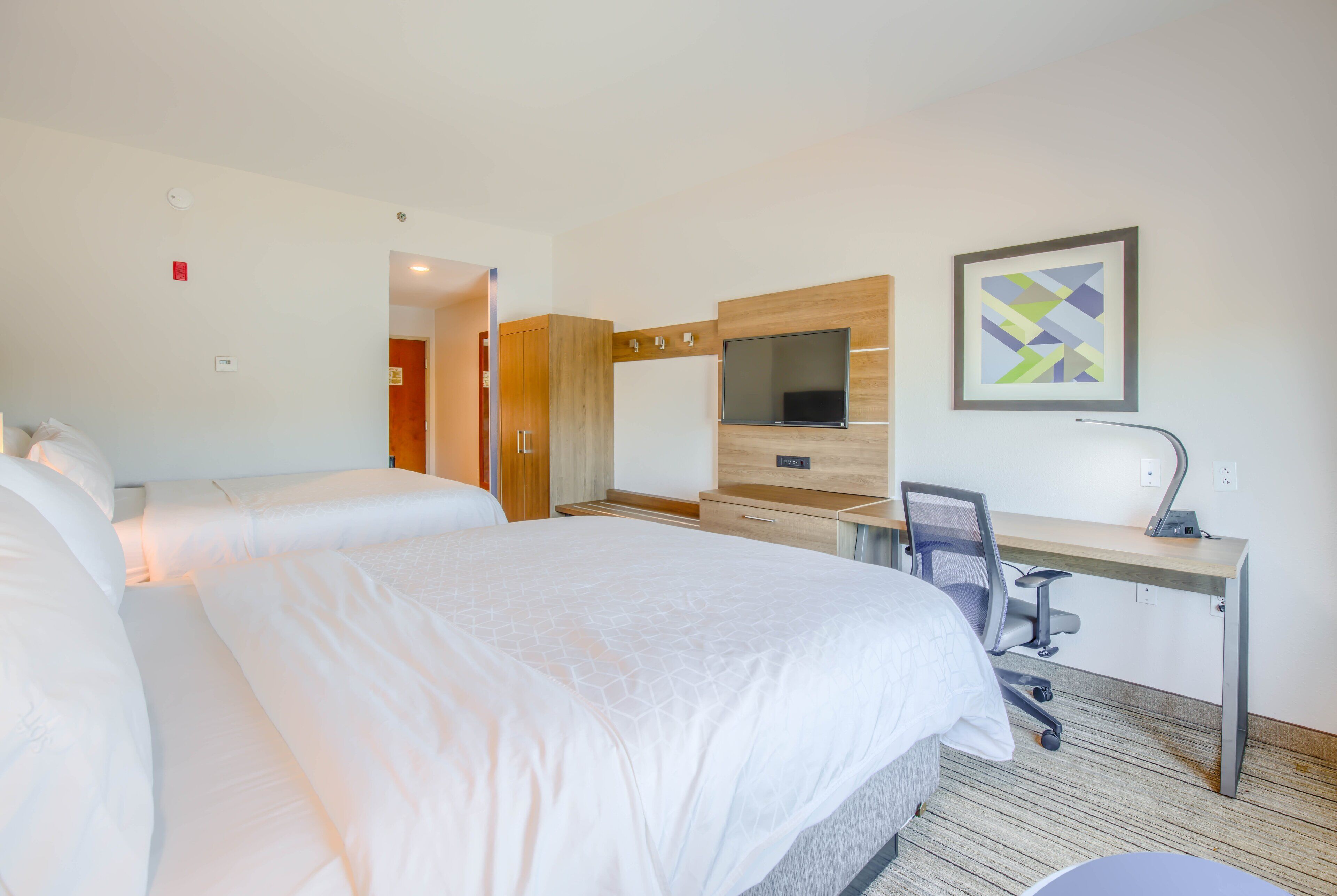 A hotel room with two beds, a desk with a chair, a TV, and a framed artwork on the wall.