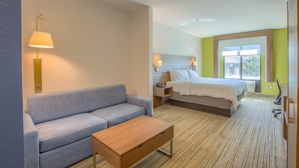 A hotel room with a sofa, a wooden table, a bed, a nightstand, and a window with a view. - Holiday Inn Express & Suites Gulf Shores by IHG (Gulf Shores)