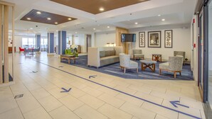A spacious lobby with a modern design, featuring a large sofa, armchairs, and a coffee table. There are directional arrows on the floor, a television mounted on the wall, and framed artwork. - Holiday Inn Express & Suites Gulf Shores by IHG (Gulf Shores)