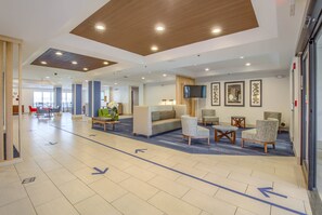 A spacious lobby with a modern design, featuring a large sofa, armchairs, and a coffee table. There are directional arrows on the floor, a television mounted on the wall, and framed artwork. - Holiday Inn Express & Suites Gulf Shores by IHG (Gulf Shores)
