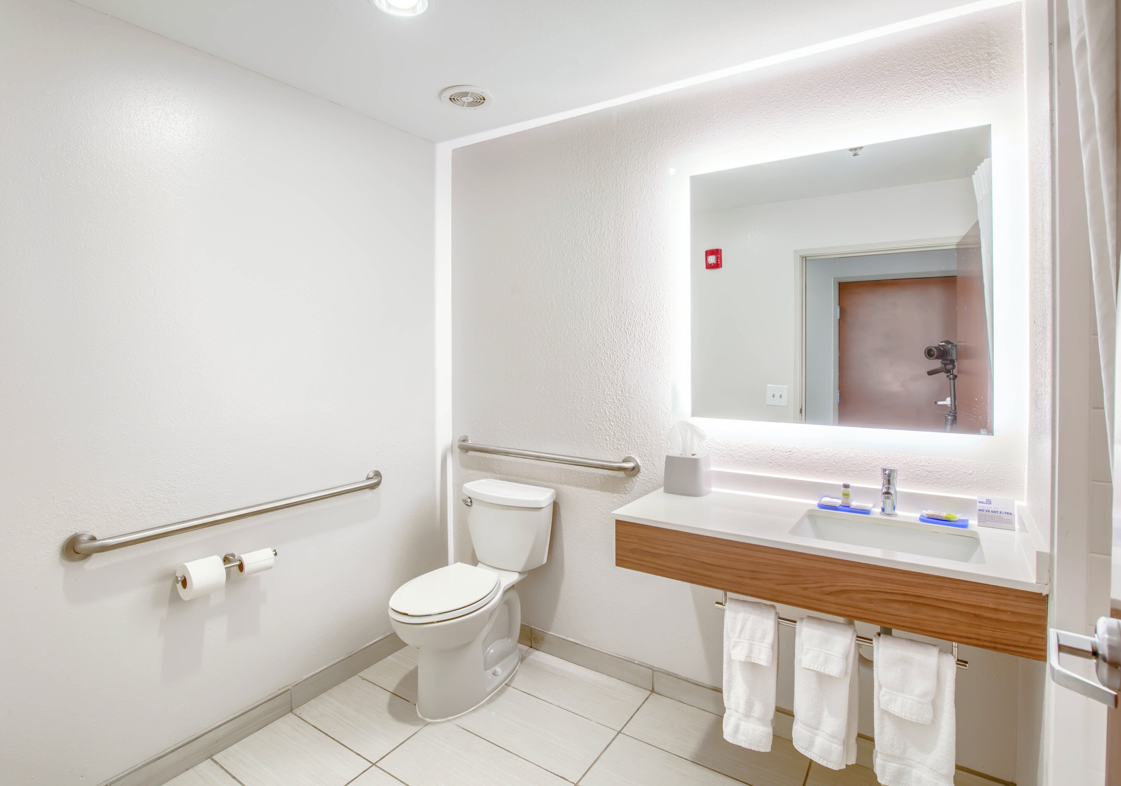 A modern bathroom with a toilet, handrails, a sink with a mirror, and a towel rack.