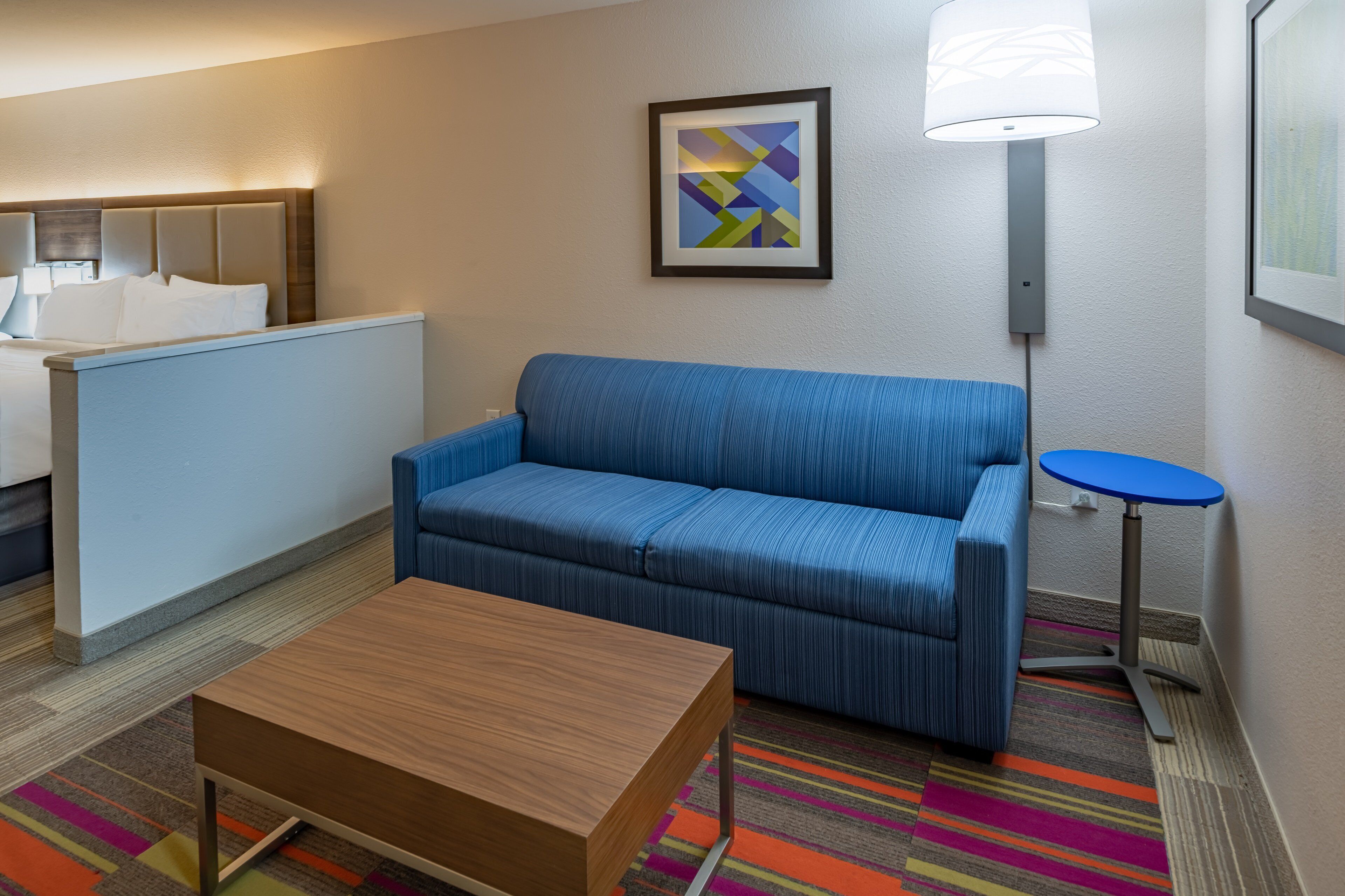 A hotel room with a blue sofa, a wooden coffee table, a small round table, a lamp, and a colorful striped rug.