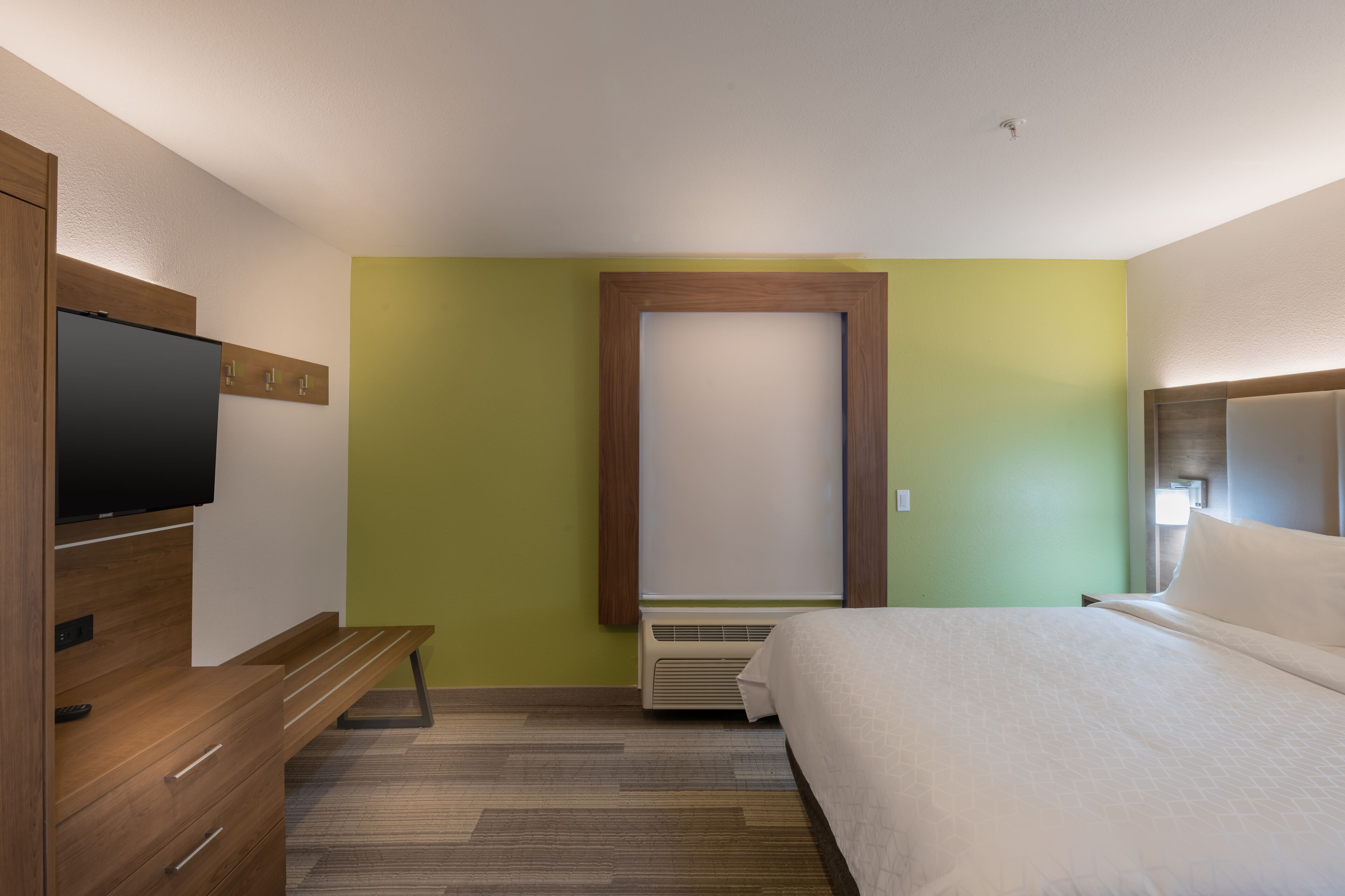A hotel room with a bed, a TV mounted on a wooden dresser, a bench, and a large mirror.