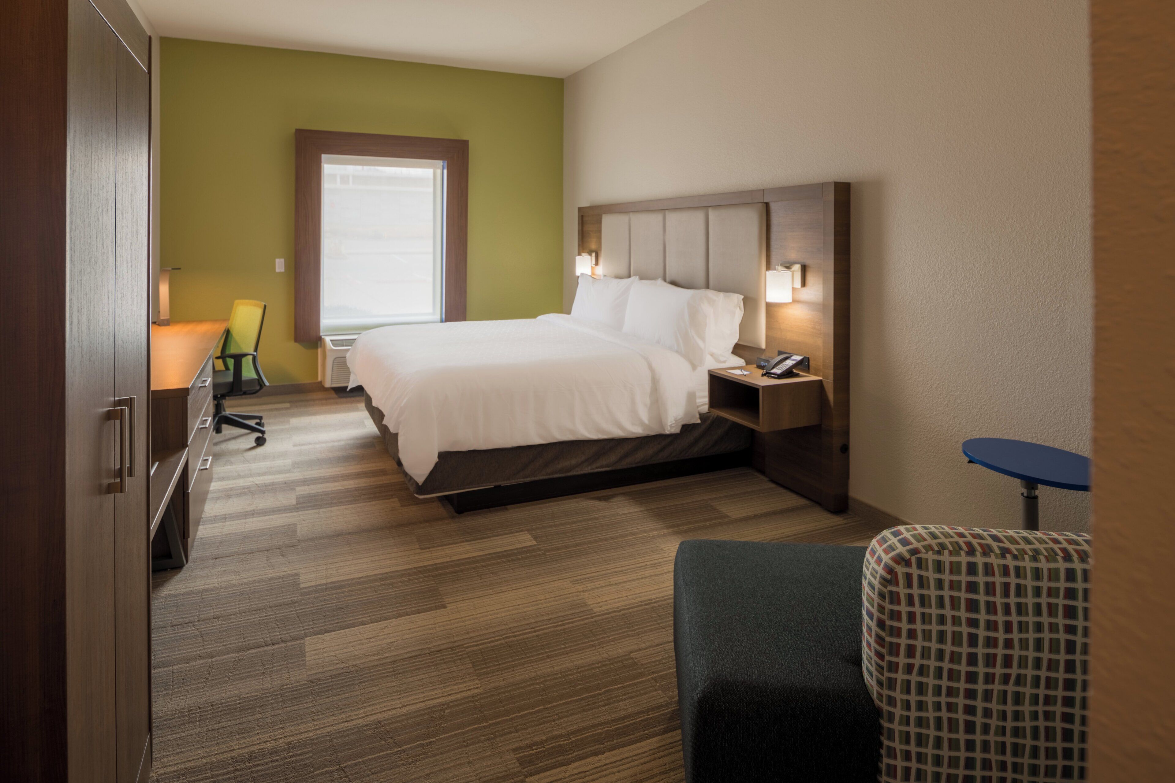 A hotel room with a bed, bedside table, lamp, chair, desk, and a window.