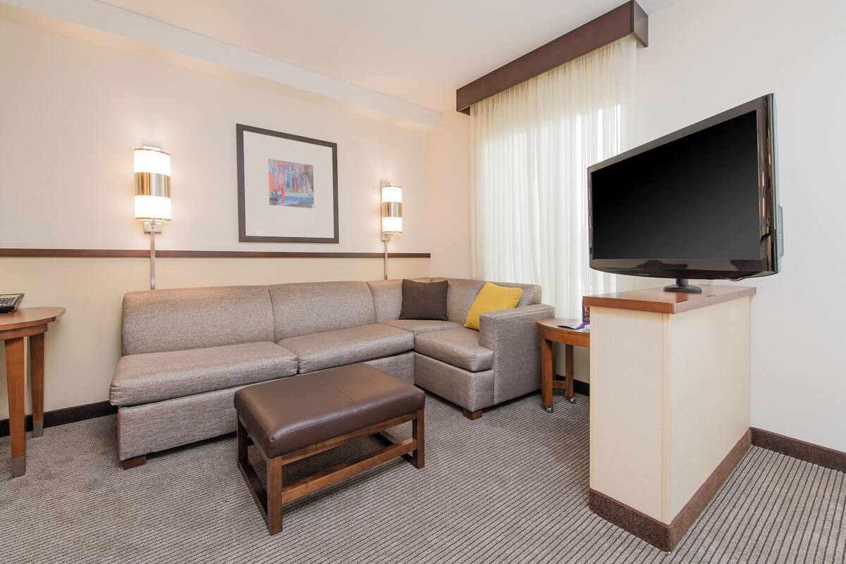 A hotel room with a sectional sofa, a flat-screen TV, a wooden coffee table, and two bedside tables with lamps.