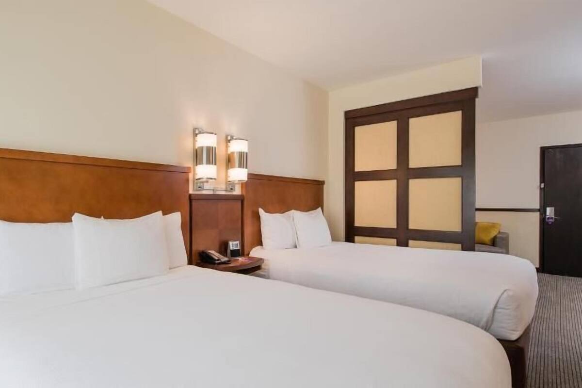A hotel room with two beds, a wooden headboard, a nightstand with a phone, and a wall-mounted light fixture.