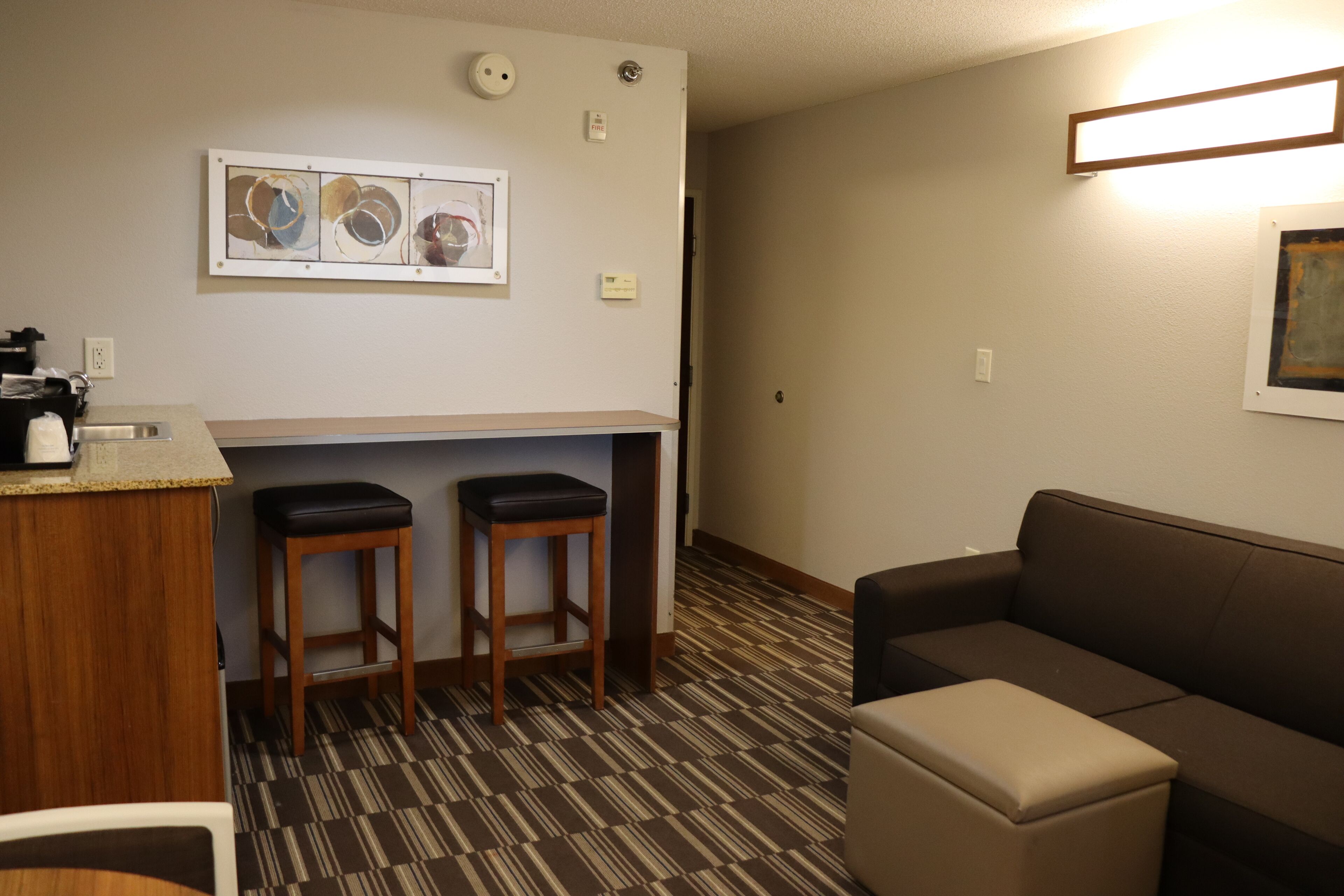 A hotel room with a kitchenette, two bar stools, a sofa, and a small ottoman.