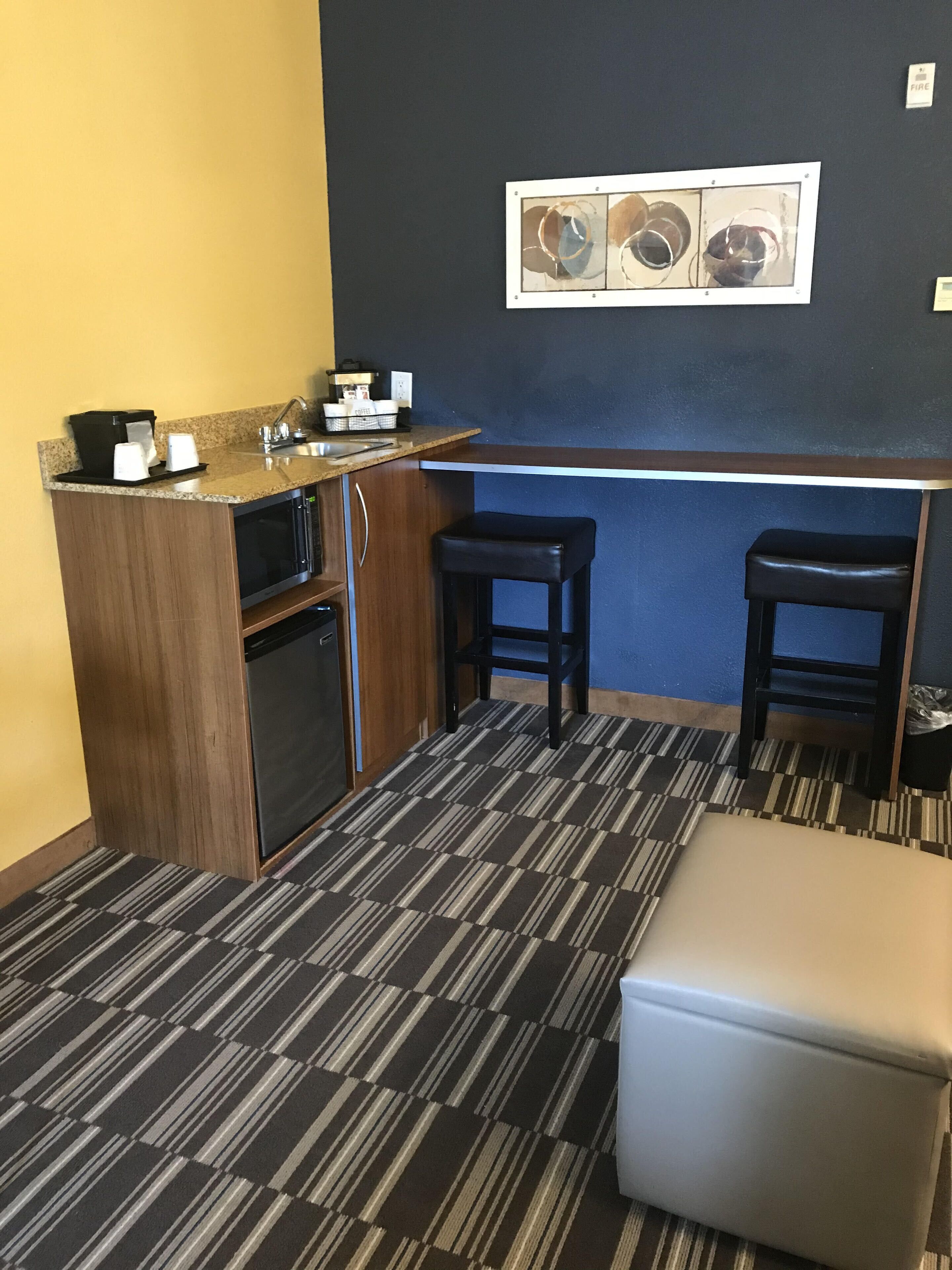 A compact kitchen with a coffee machine, sink, and a microwave. There are two stools and a small table. The floor has a striped carpet.