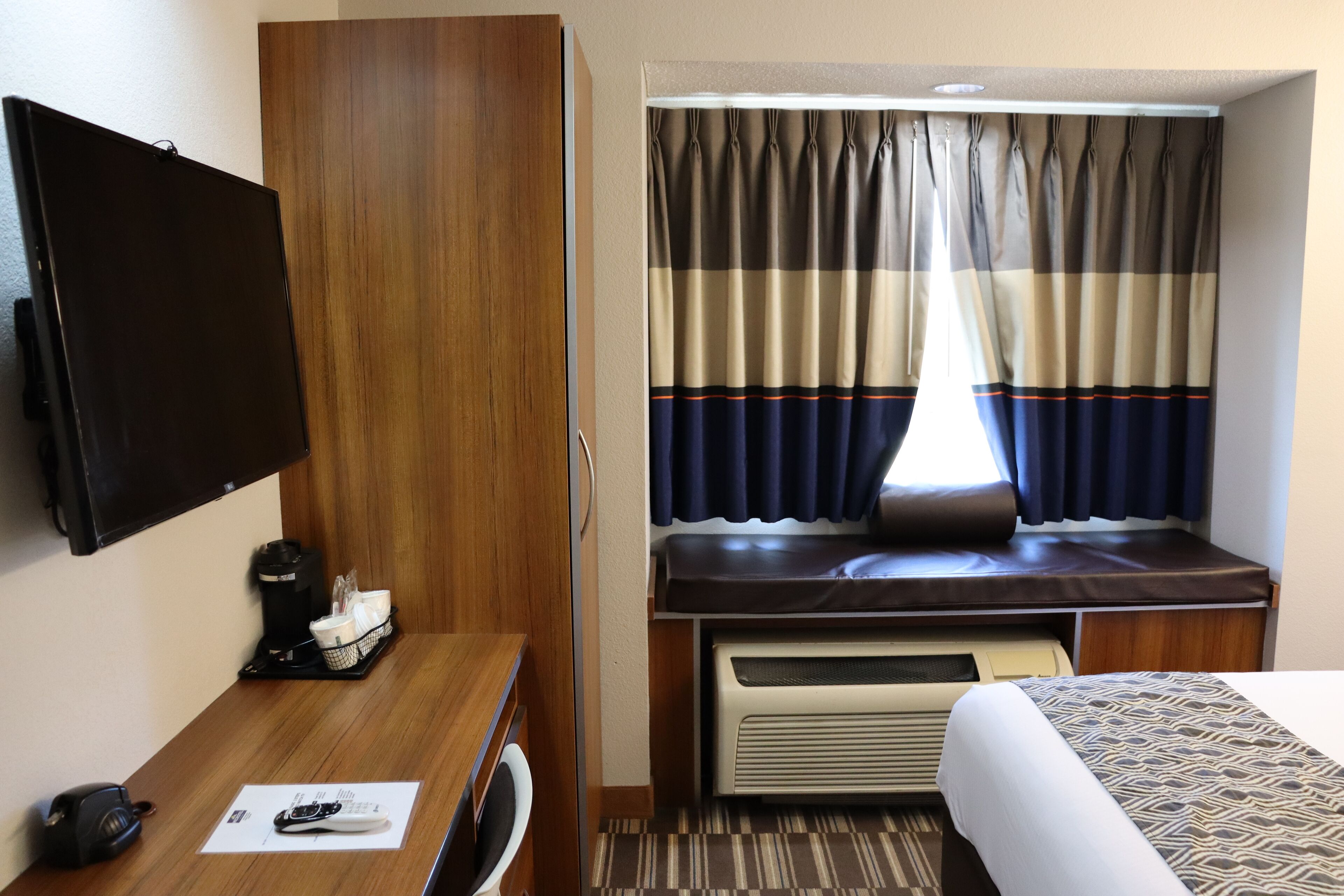 A compact hotel room with a bed, a desk with a coffee maker, a television, and a window with curtains.