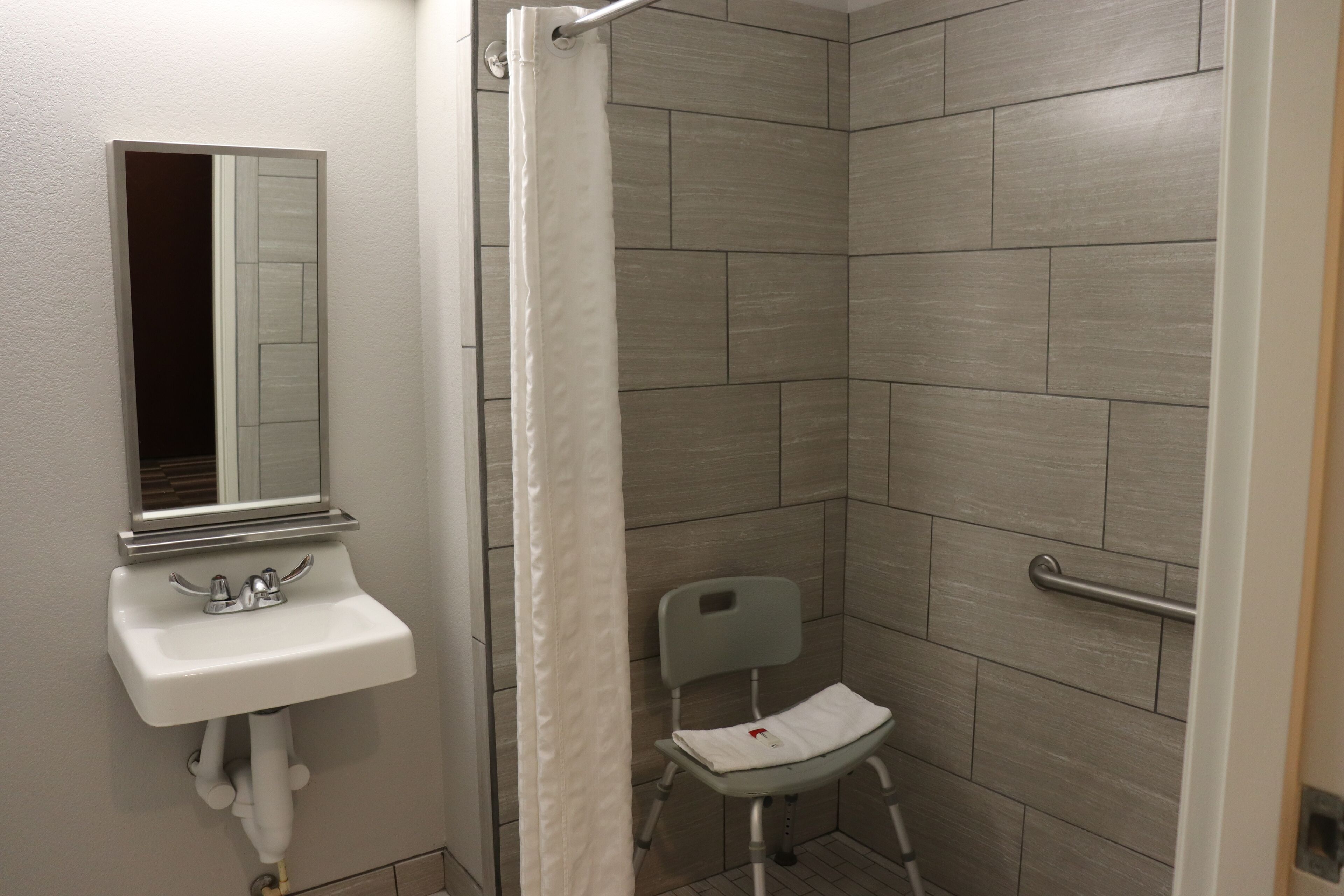 A small bathroom with a white sink, a mirror, a shower curtain, and a wheelchair-accessible chair.