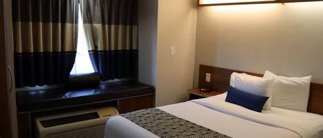 Una habitación de hotel con una cama, un escritorio, una silla, un ventanal con cortinas y una lámpara fijada a la pared.