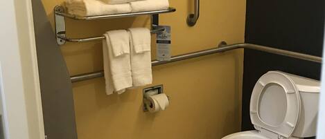 A small bathroom with a toilet, a handrail, a towel rack with towels, and a toilet paper dispenser.
