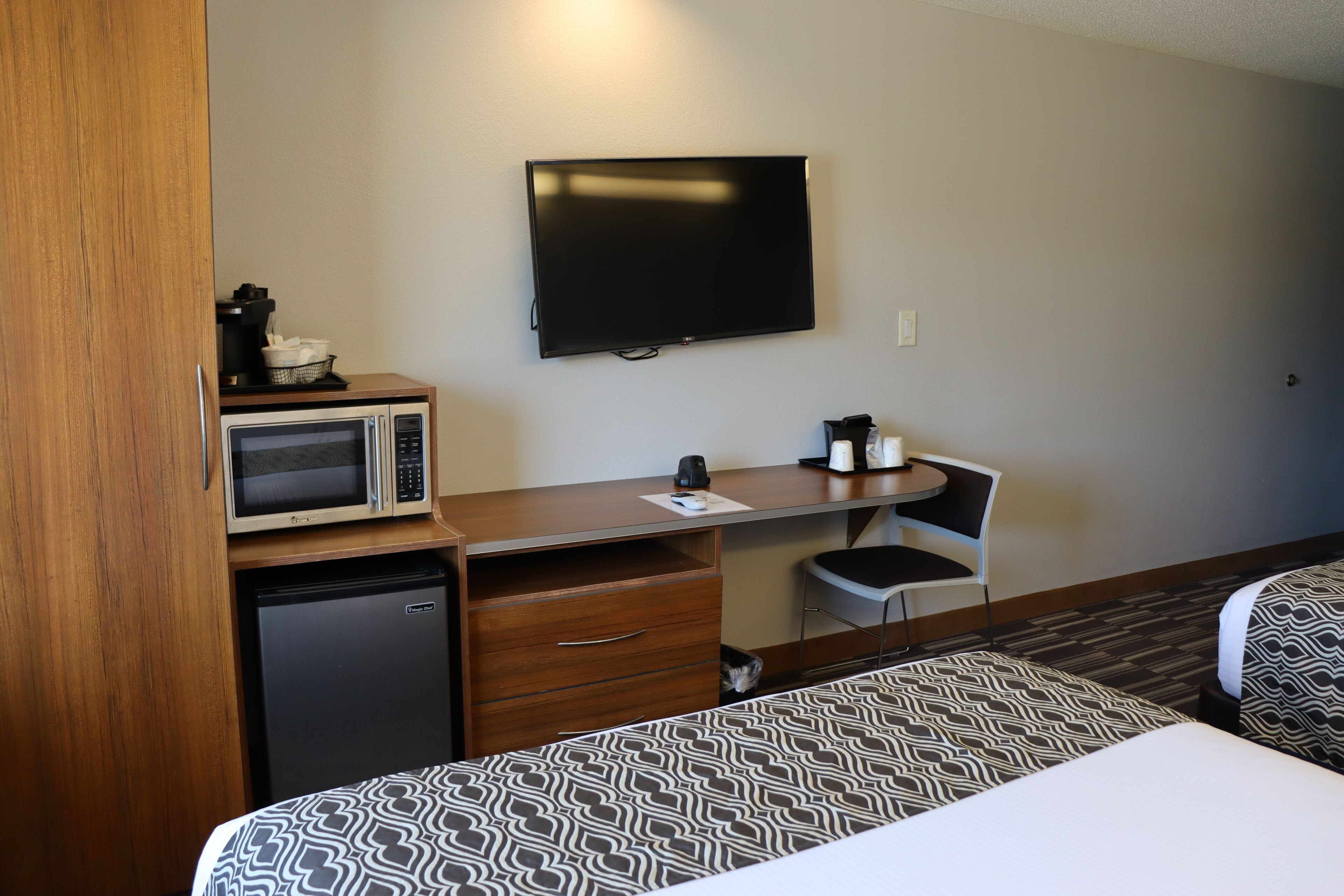 A hotel room with a bed, a desk with a microwave, a small refrigerator, a television, and a chair.