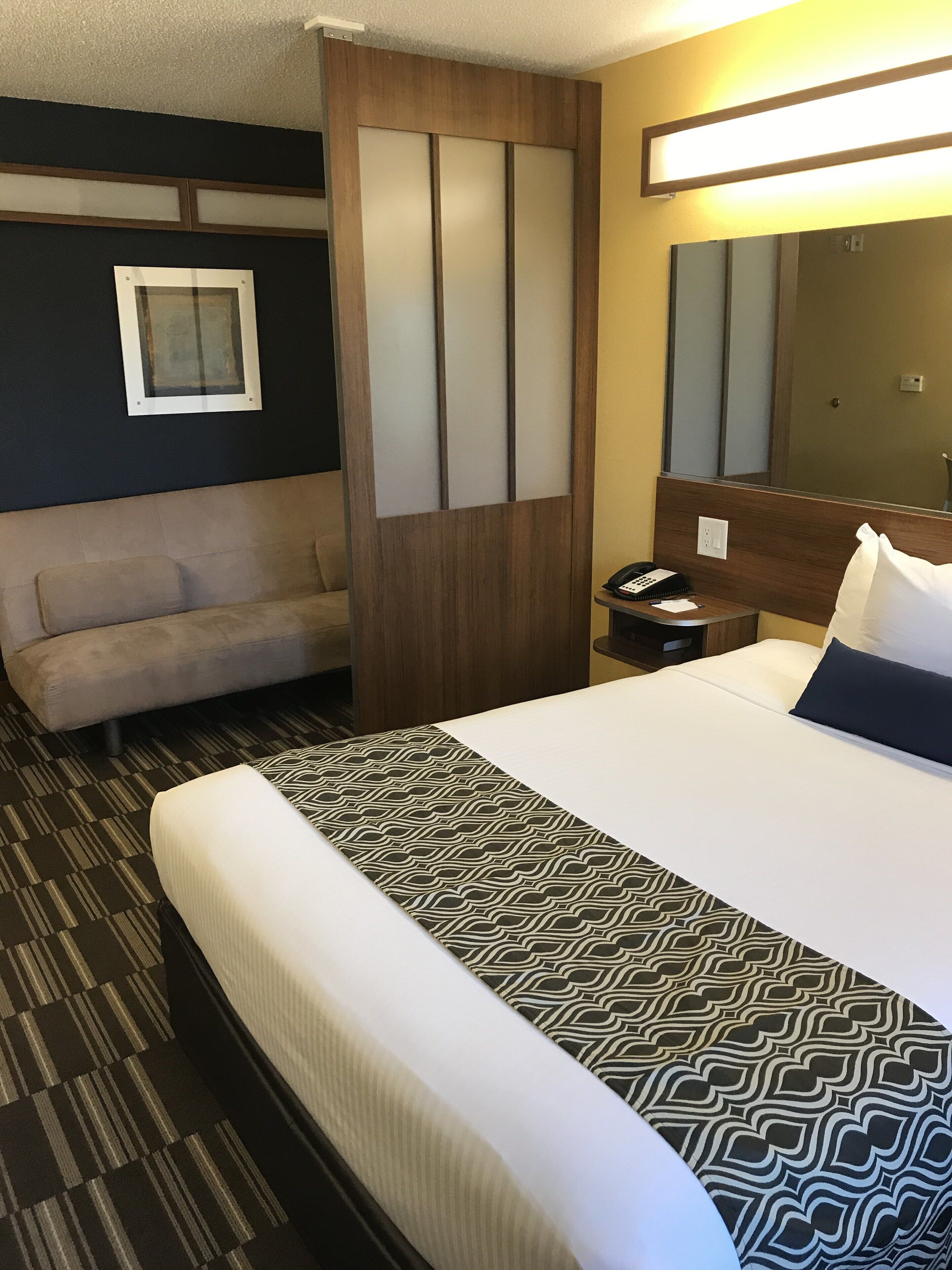 A hotel room with a bed, a sofa, a desk with a phone, a mirror, and a wall-mounted light fixture.