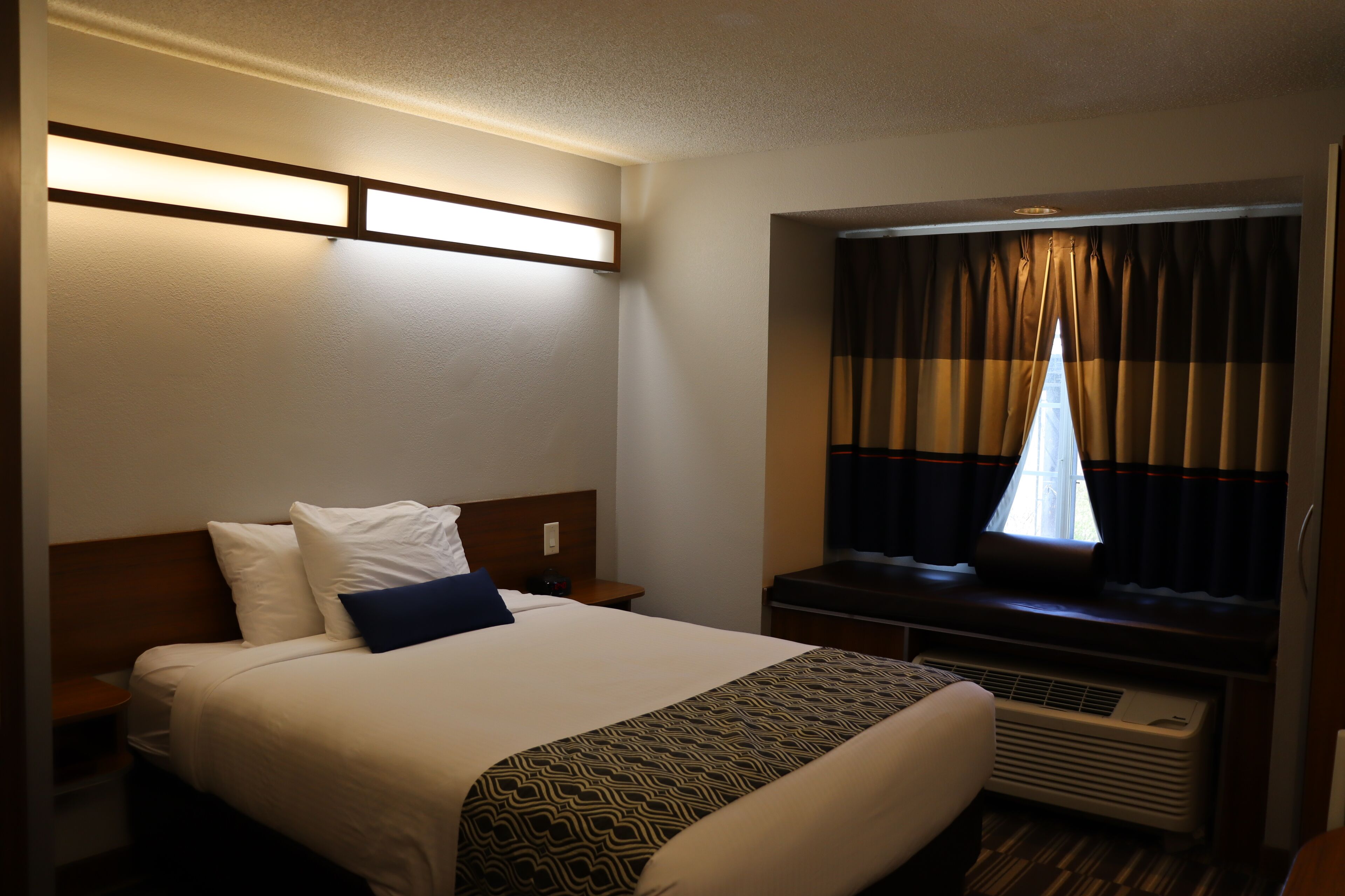 A hotel room with a large bed, a bedside table, a window with curtains, and a wall-mounted air conditioning unit.