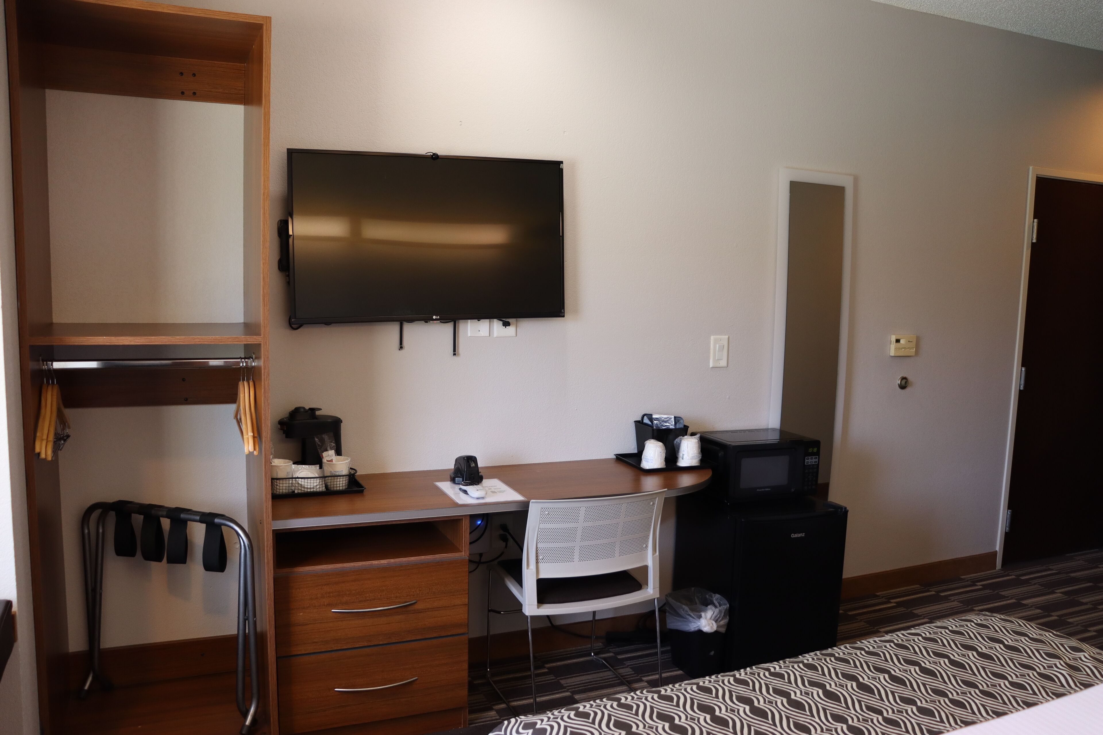 A hotel room with a wooden desk, a white chair, a television mounted on the wall, a microwave, a small refrigerator, a desk lamp, and a patterned rug.