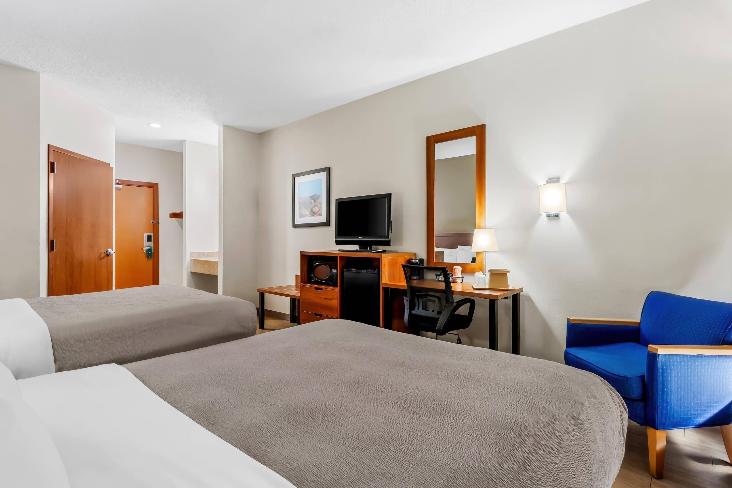 Habitación de hotel con dos camas, un escritorio con computadora, una silla, televisor y espejo.