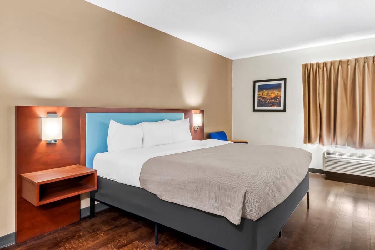 Habitación de hotel con cama, mesitas de noche, lámpara de pared, ventana con cortinas e imagen enmarcada en la pared.