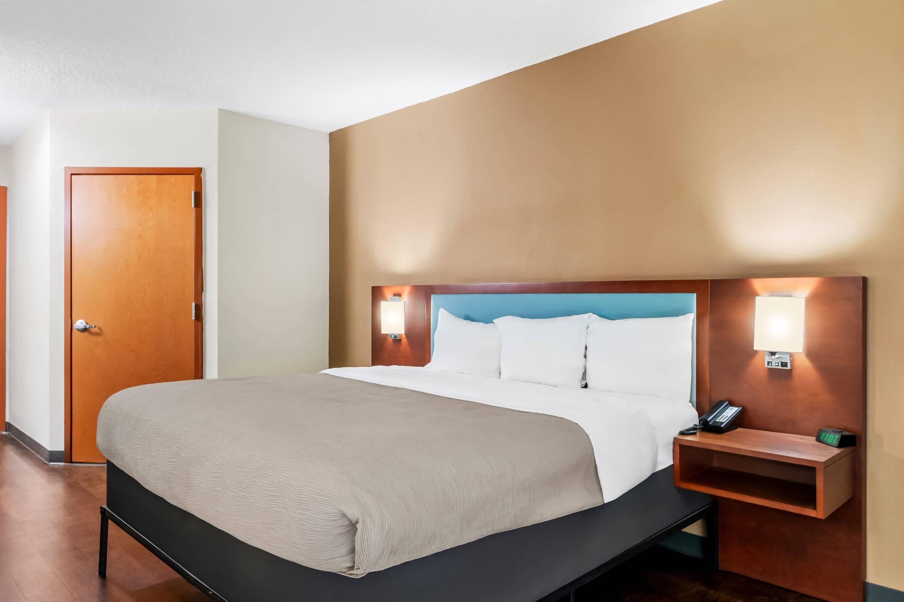 A hotel room with a bed, bedside table, lamp, and a door.