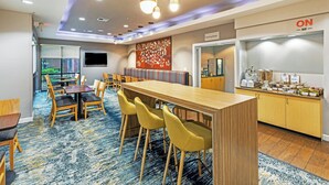 Een moderne hotel lobby met een lange houten receptiebalie, gele stoelen, een grote tv aan de muur en een ontbijtbuffet met diverse producten.