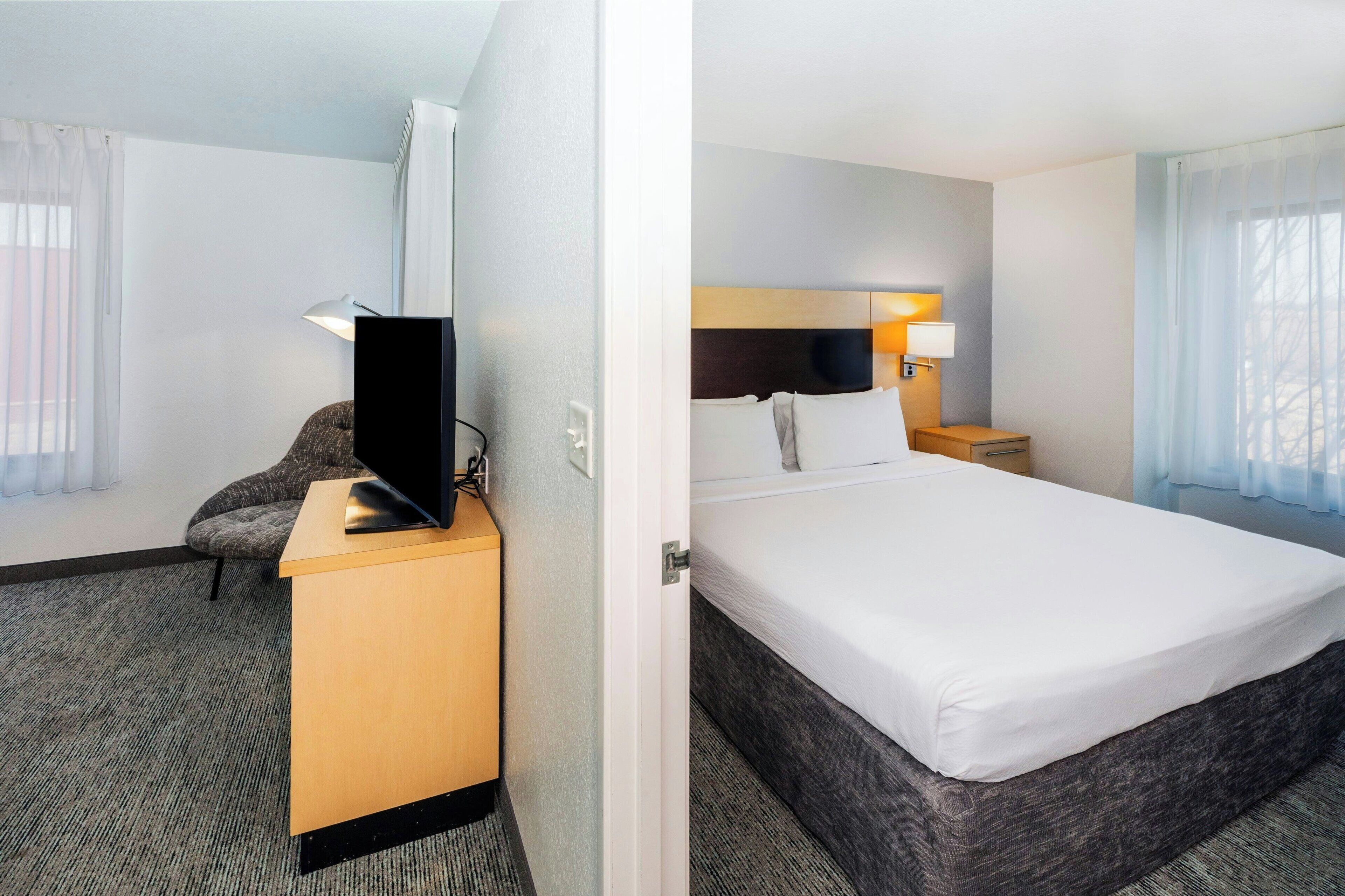A hotel room with a bed, a desk with a flat-screen TV, a chair, and a nightstand with a lamp.