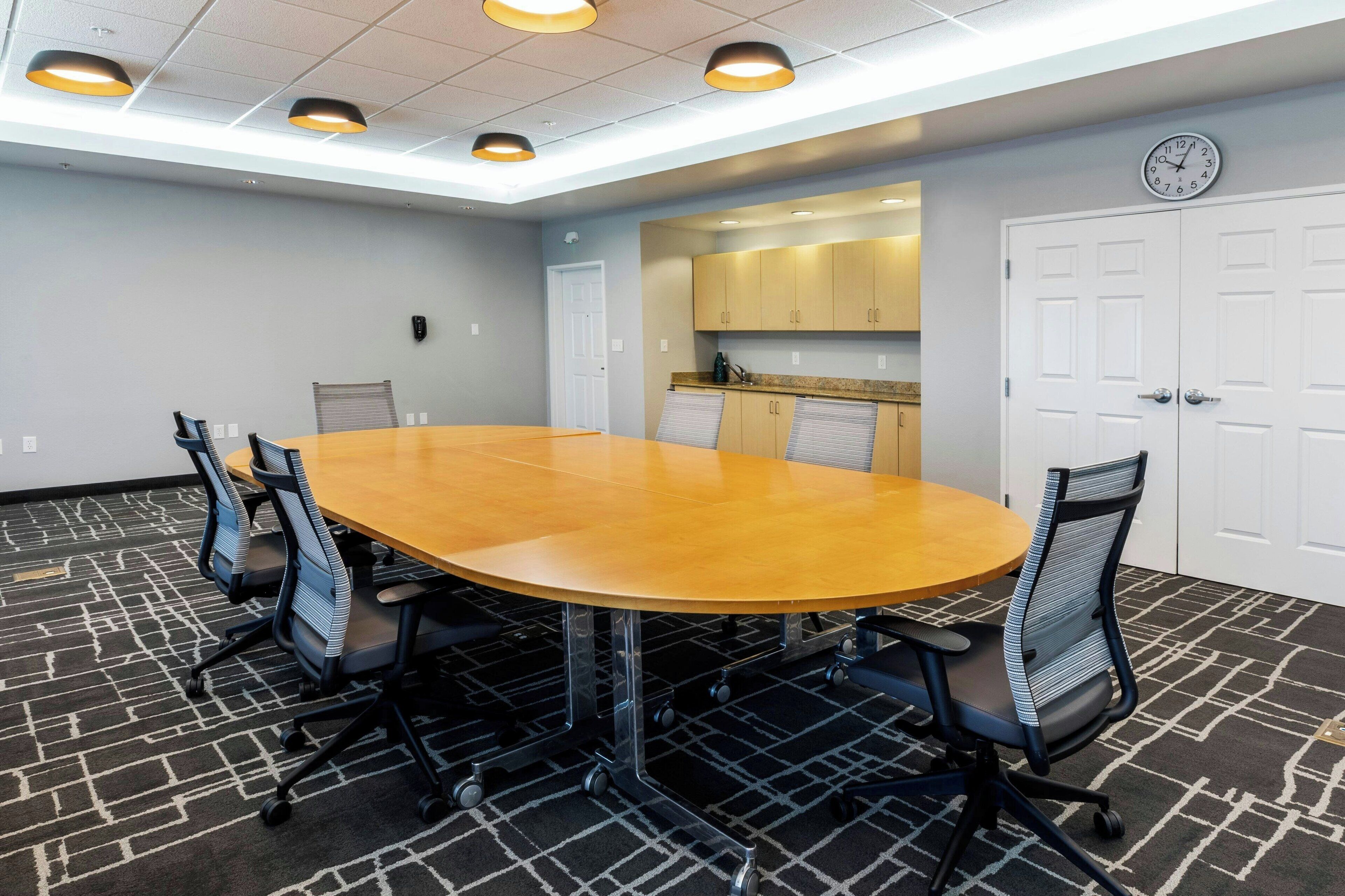 A conference room with a large oval table, six office chairs, a clock on the wall, and a kitchenette in the background.
