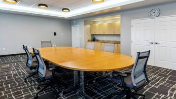 A conference room with a large oval table, six office chairs, a clock on the wall, and a kitchenette in the background.