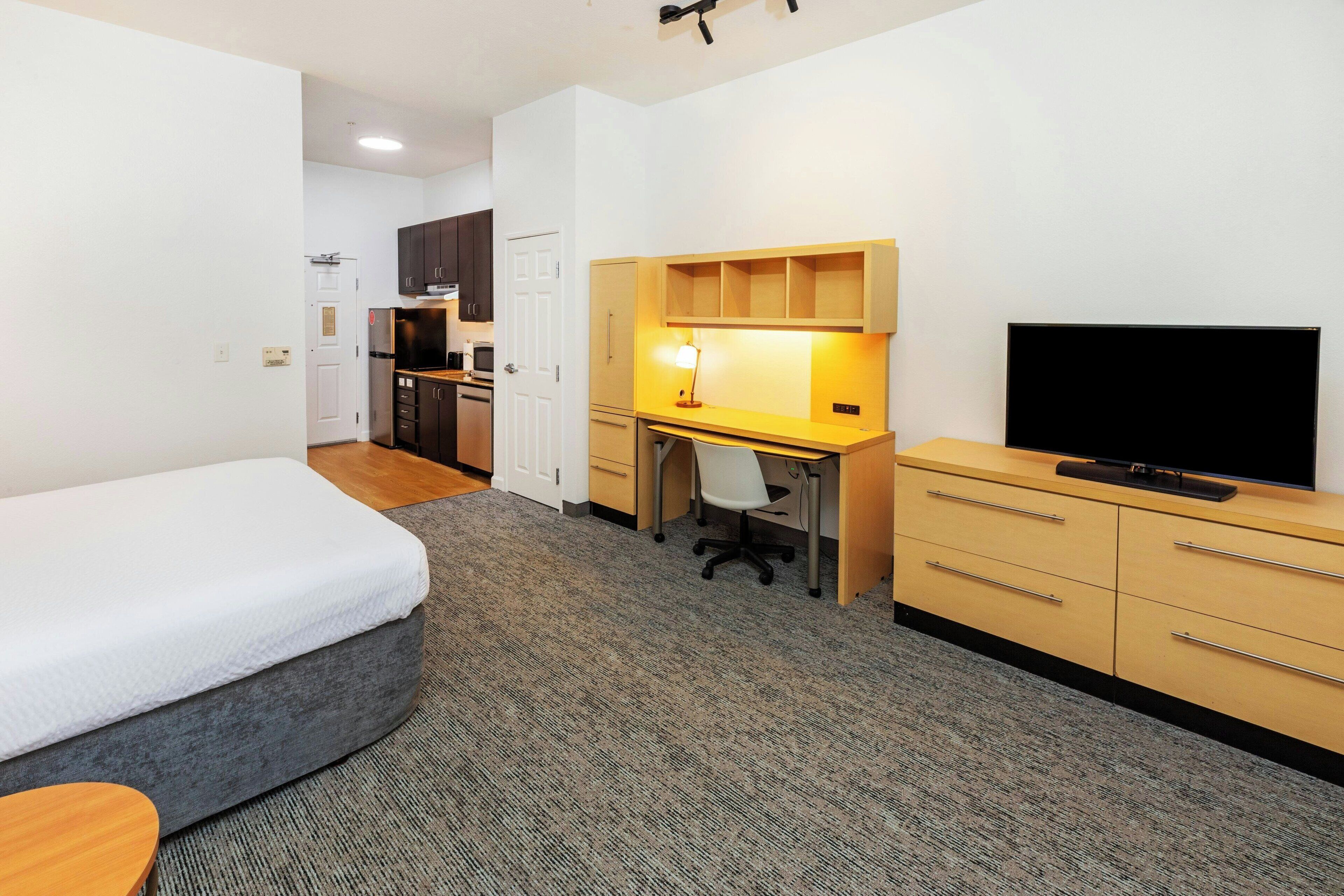 A compact hotel room with a bed, a desk, a TV, and a kitchenette.