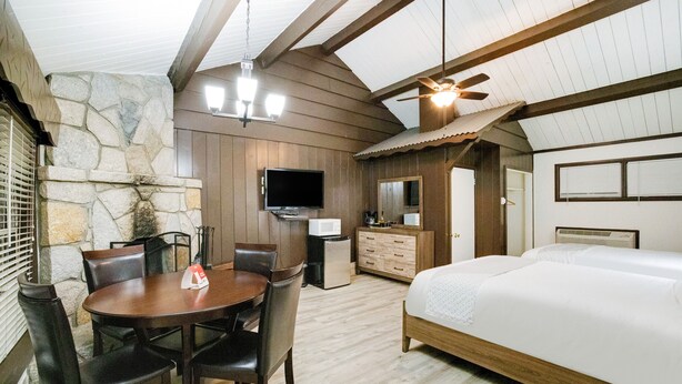 A room with a bed, a TV, a dining table with chairs, and a stone wall. - Arrowhead Tree Top Lodge (Lake Arrowhead)
