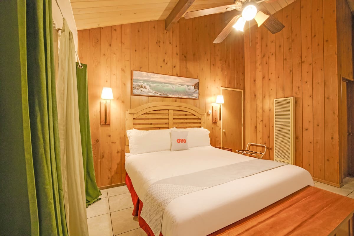 Une chambre à coucher aux murs lambrissés, avec un lit, deux lampes fixées au mur, un ventilateur de plafond et un tableau représentant un paysage.