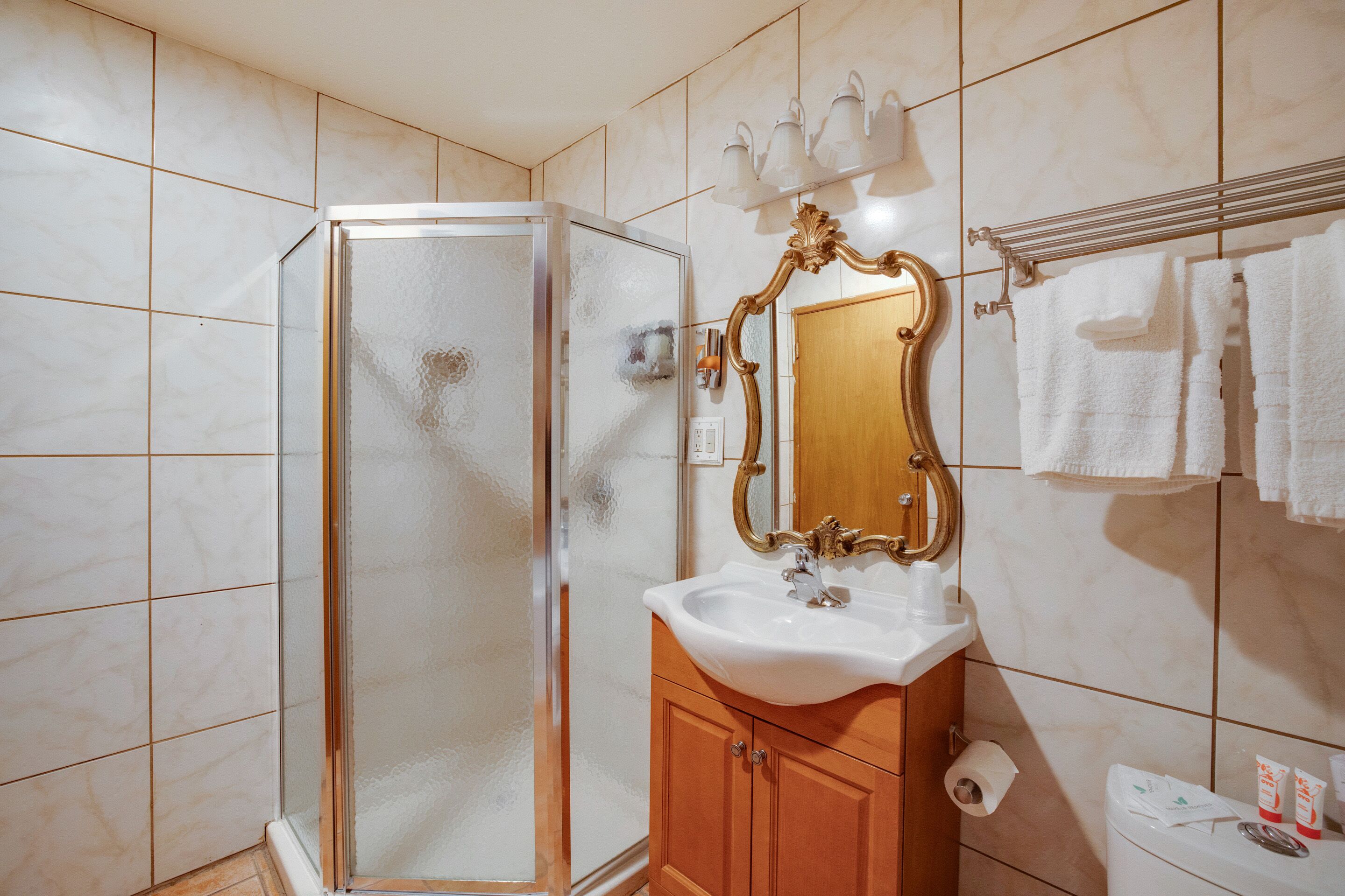 A bathroom with a glass-enclosed shower, a white sink, a wooden vanity, a mirror with an ornate frame, and a towel rack with white towels.