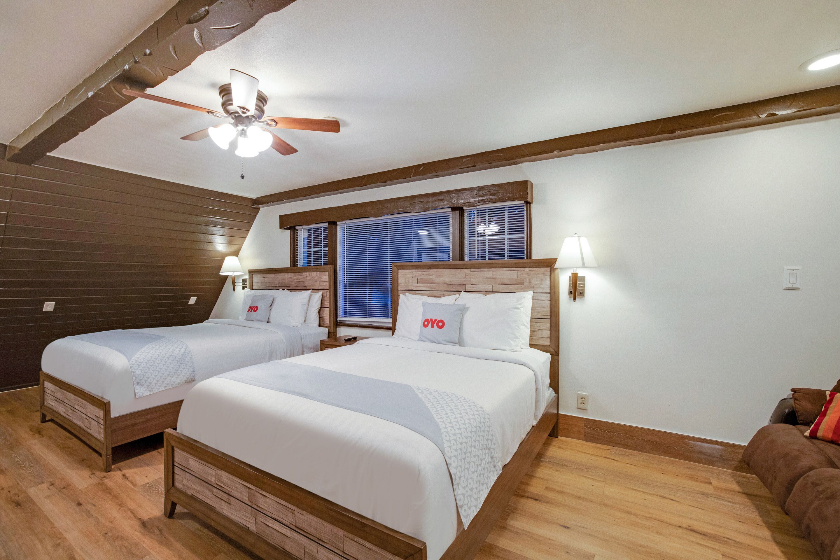 A hotel room with two beds, wooden floors, a ceiling fan, and a window with blinds.