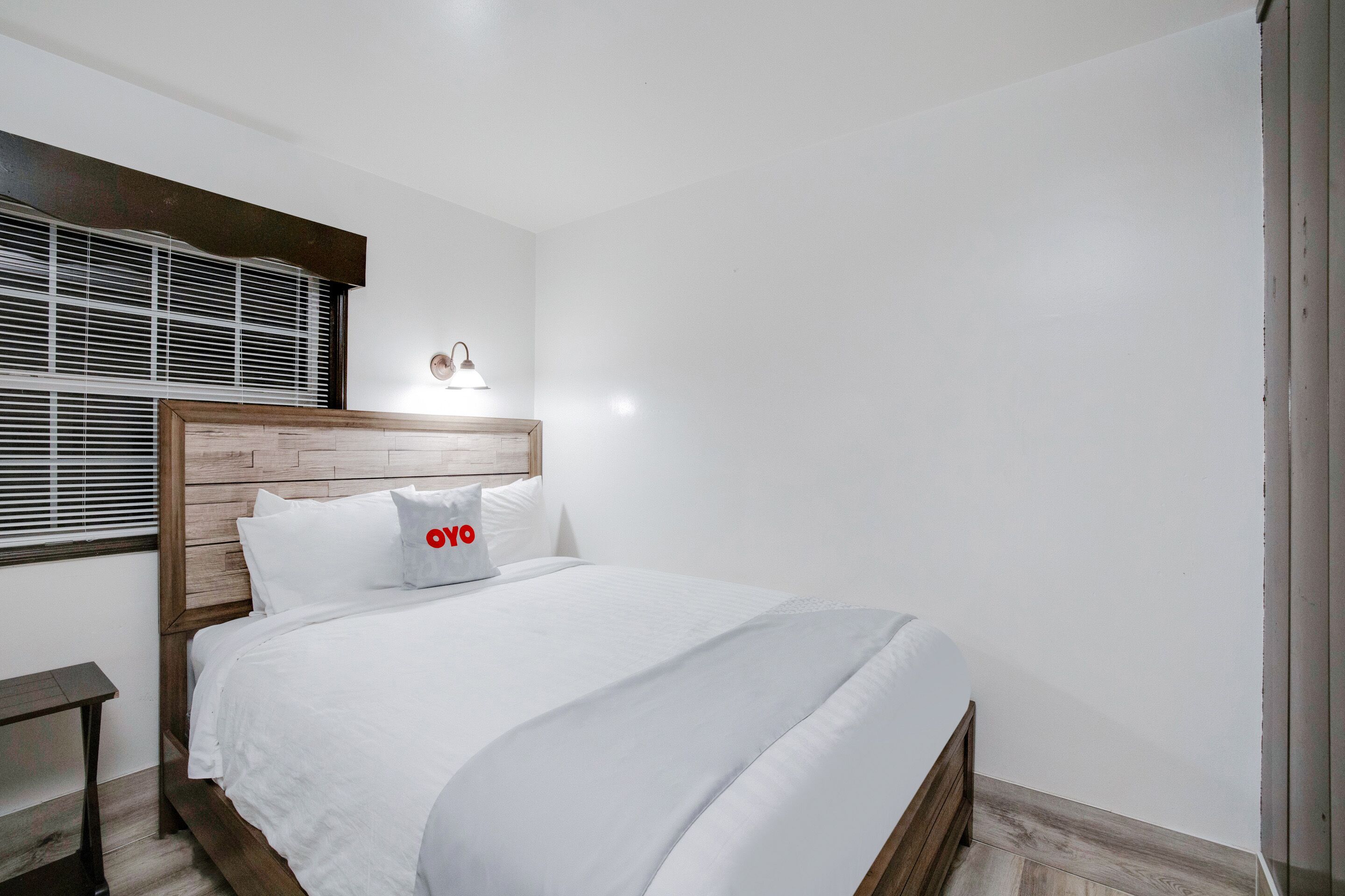 A neatly made bed with white bedding and a wooden headboard, a bedside table, and a window with blinds.