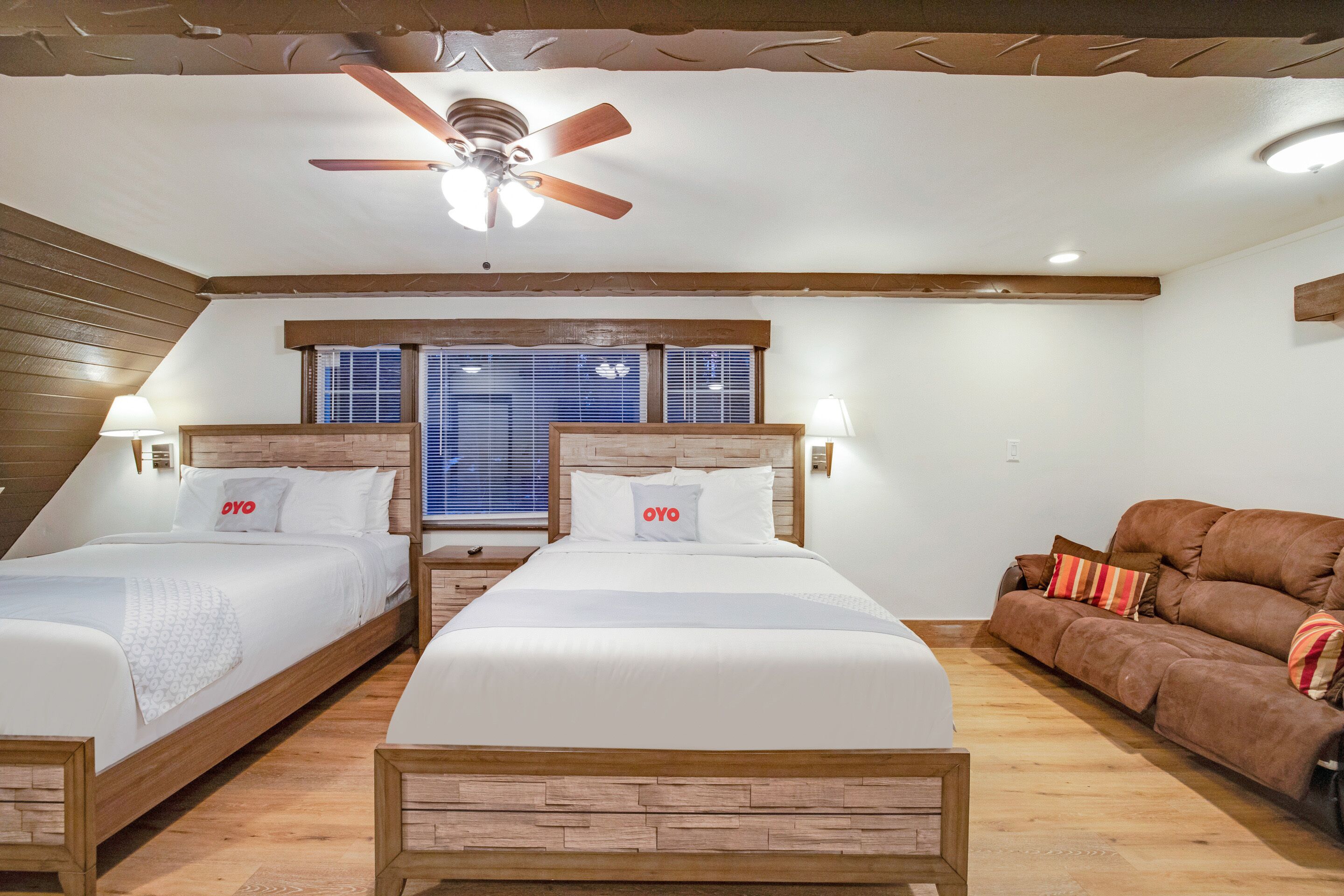 A bedroom with two beds, a ceiling fan, wooden beams, a window with blinds, and a brown sofa.