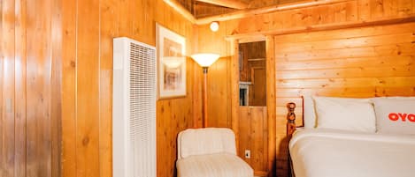 A wooden cabin room with a bed, a chair, a heater, and a wall-mounted lamp.