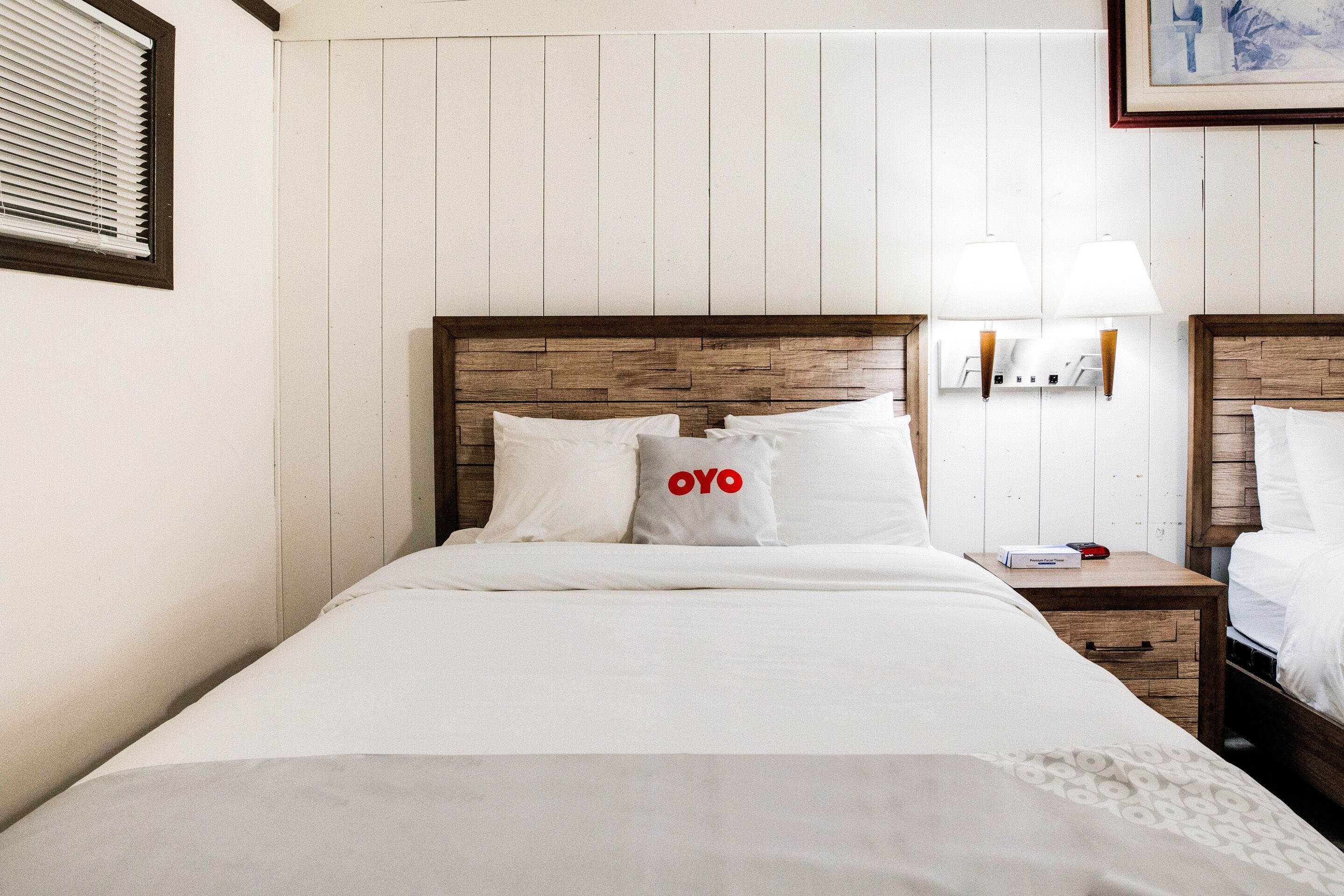A hotel room with a wooden headboard, white bedding, a bedside table with a lamp, and a framed picture on the wall.