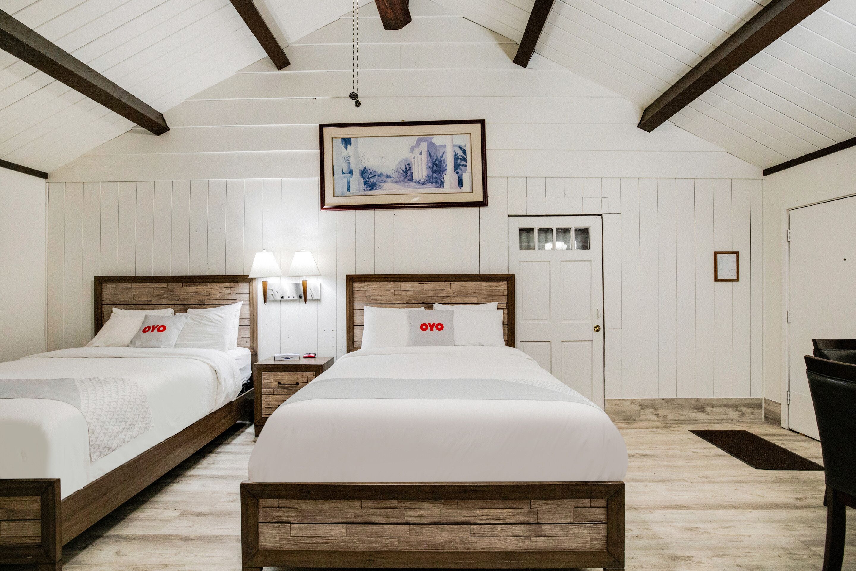A bedroom with two beds, wooden headboards, white walls, a framed picture on the ceiling, and a door in the background.