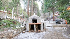 A stone-built outdoor fireplace surrounded by a stone wall and wooden steps, with a forested backdrop and a building with a white fence in the background. - Arrowhead Tree Top Lodge (Lake Arrowhead)