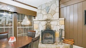A stone fireplace with a metal screen, a dining table with chairs, and a window with blinds. - Arrowhead Tree Top Lodge (Lake Arrowhead)