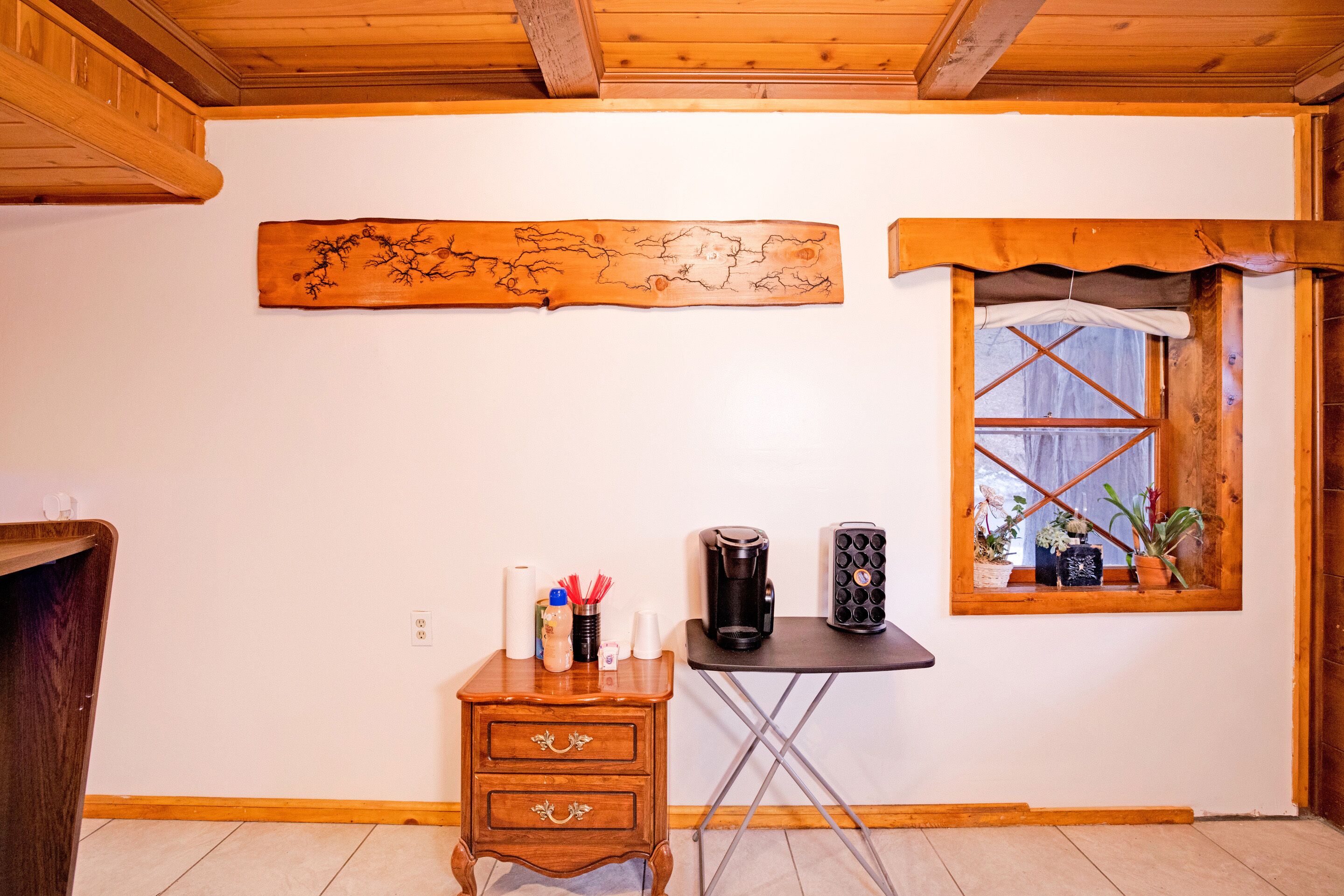 Una stanza con soffitto in legno, un'opera d'arte appesa al muro, un piccolo comò di legno, una macchina da caffè e una finestra con piante.