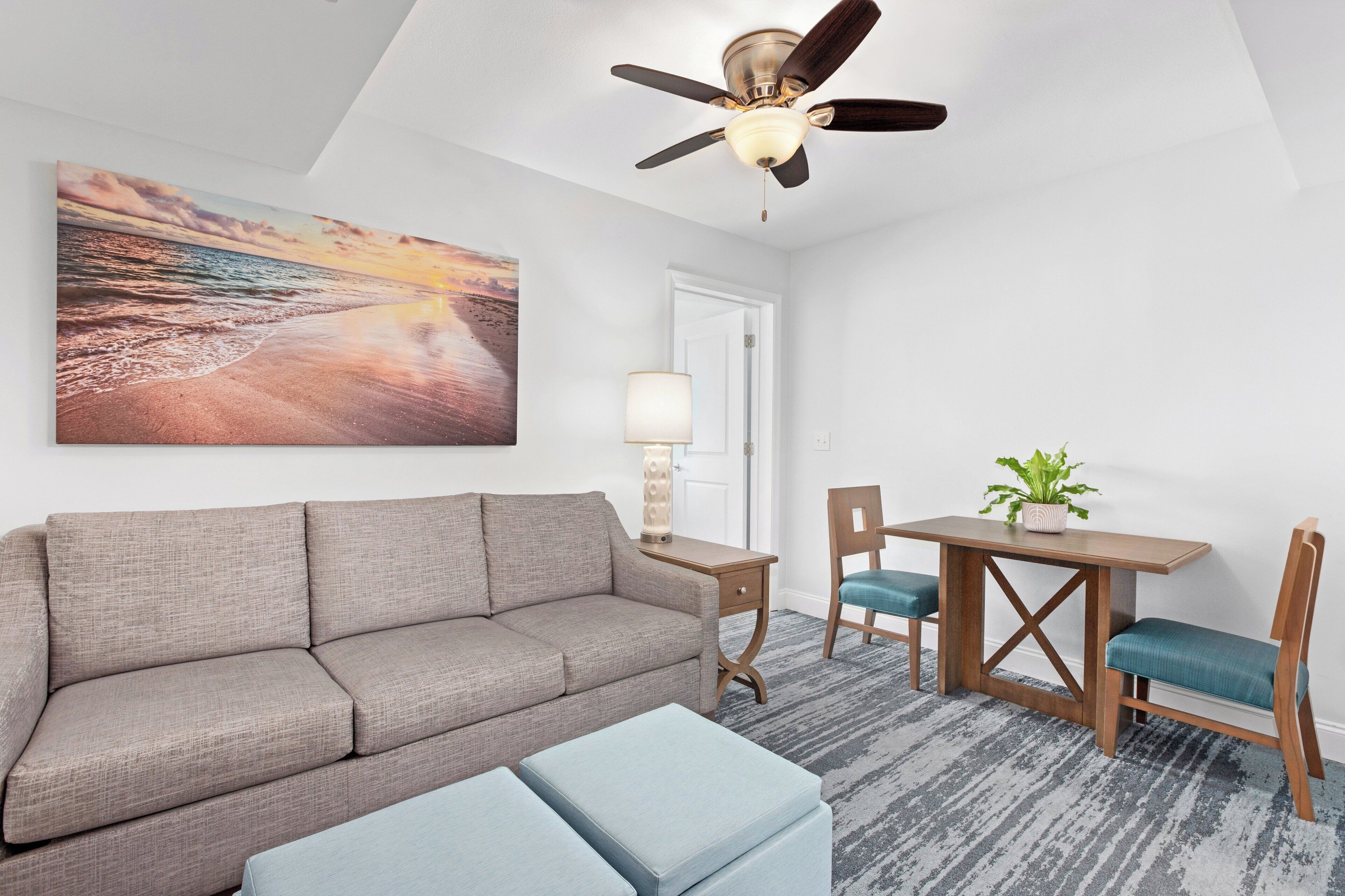 A room with a sofa, a small table with chairs, a ceiling fan, and a painting of a beach scene.