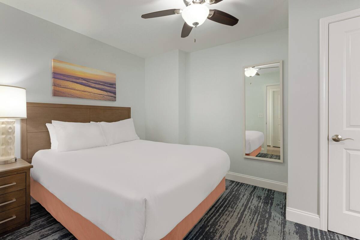 A hotel room with a bed, a ceiling fan, a nightstand with a lamp, a painting on the wall, and a mirror.